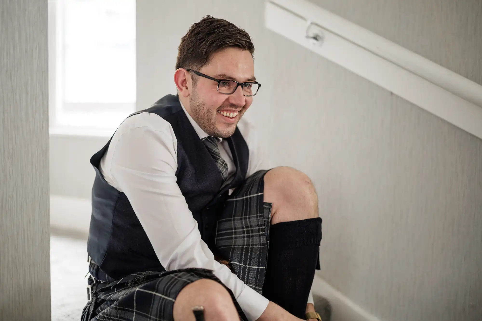 A smiling man with glasses and dark hair, wearing a white shirt, tie, dark waistcoat, and a traditional tartan kilt, sits on a carpeted staircase indoors, adjusting his black socks. Soft natural light enters through a window in the background.