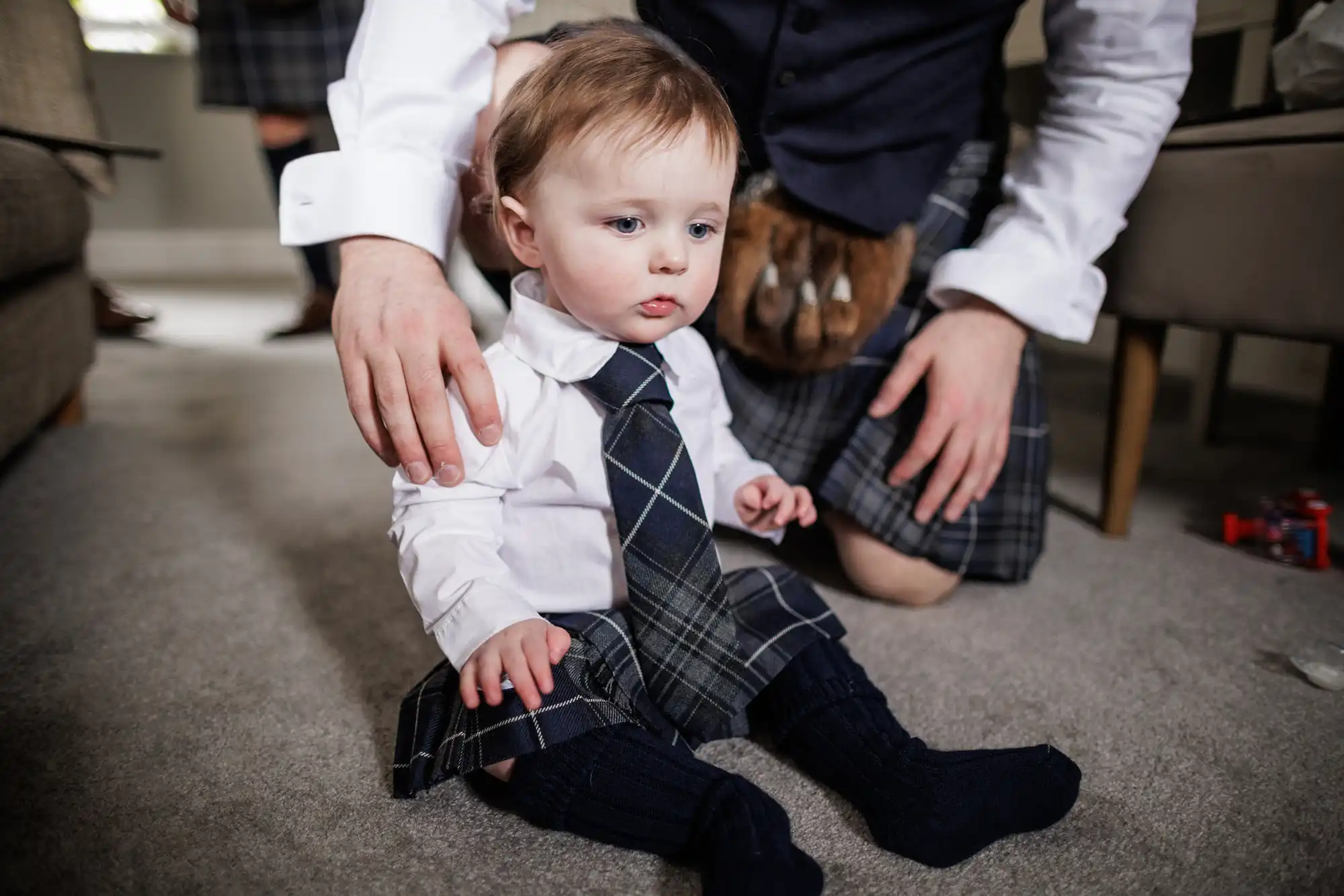 A young child sits on a carpeted floor wearing a white shirt, blue checked tie, matching kilt, and dark socks. An adult, also dressed in a kilt, is kneeling beside the child with a supportive hand on their shoulder.