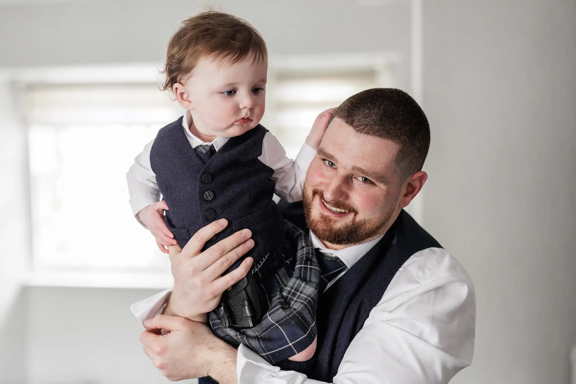 A smiling man with short hair and a beard holds a young boy. Both wear white shirts, dark waistcoats, and tartan kilts. The man looks at the camera whilst the boy looks away. They stand in a bright room with a blurred window in the background.