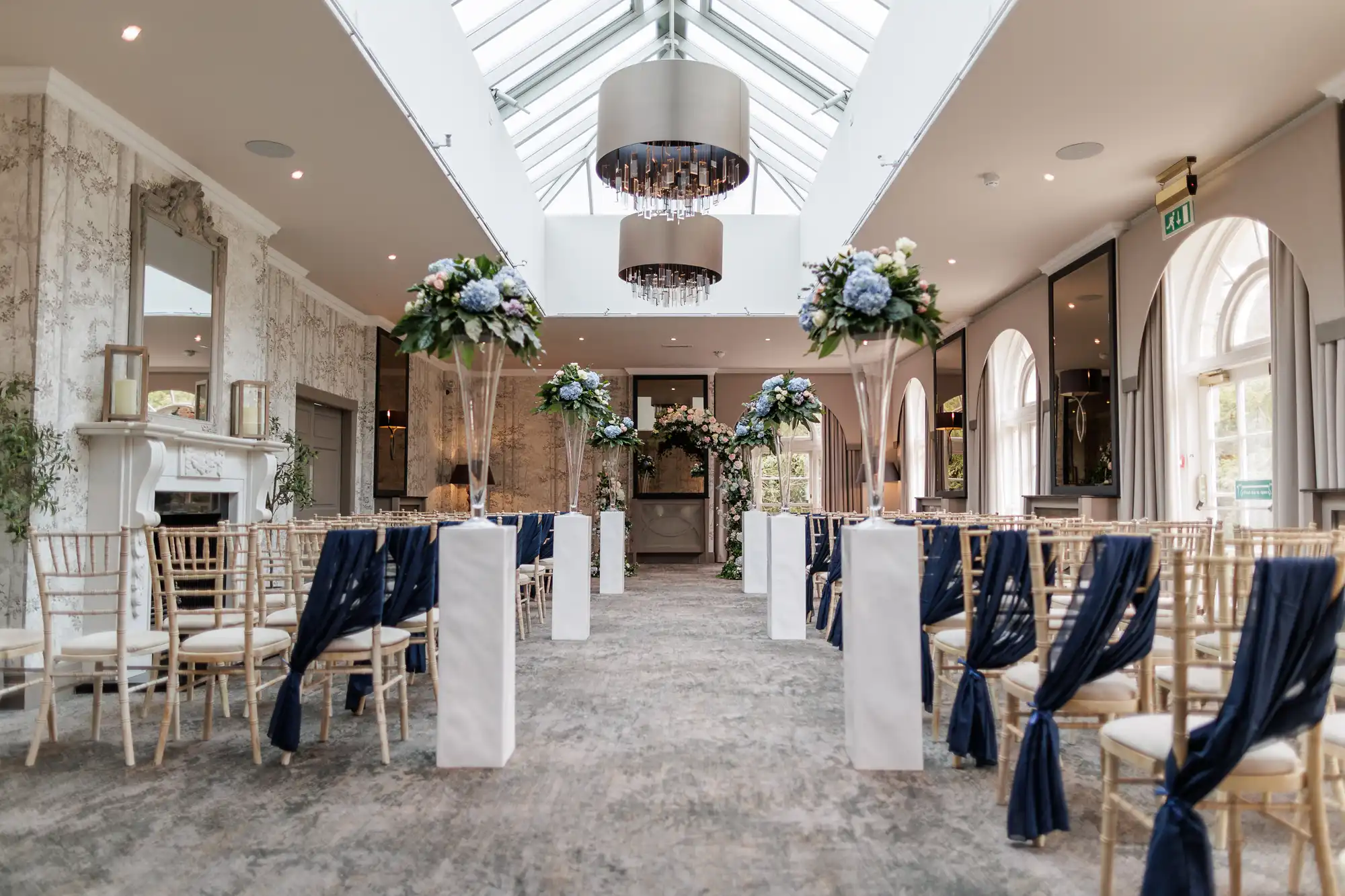 Elegant indoor wedding ceremony set-up with gold chairs draped in navy fabric, tall floral arrangements with white and blue flowers on white columns, under a glass ceiling with modern chandeliers. A white fireplace and large windows line the bright, spacious room.