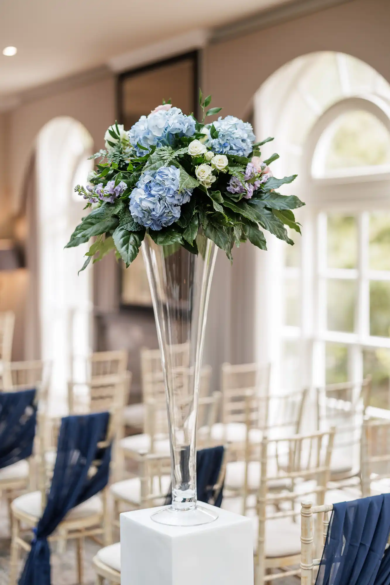 A tall, clear glass vase holds a large floral arrangement of blue hydrangeas, white roses, and greenery. The vase is placed on a white plinth in a bright room with arched windows and rows of chairs draped with navy fabric.