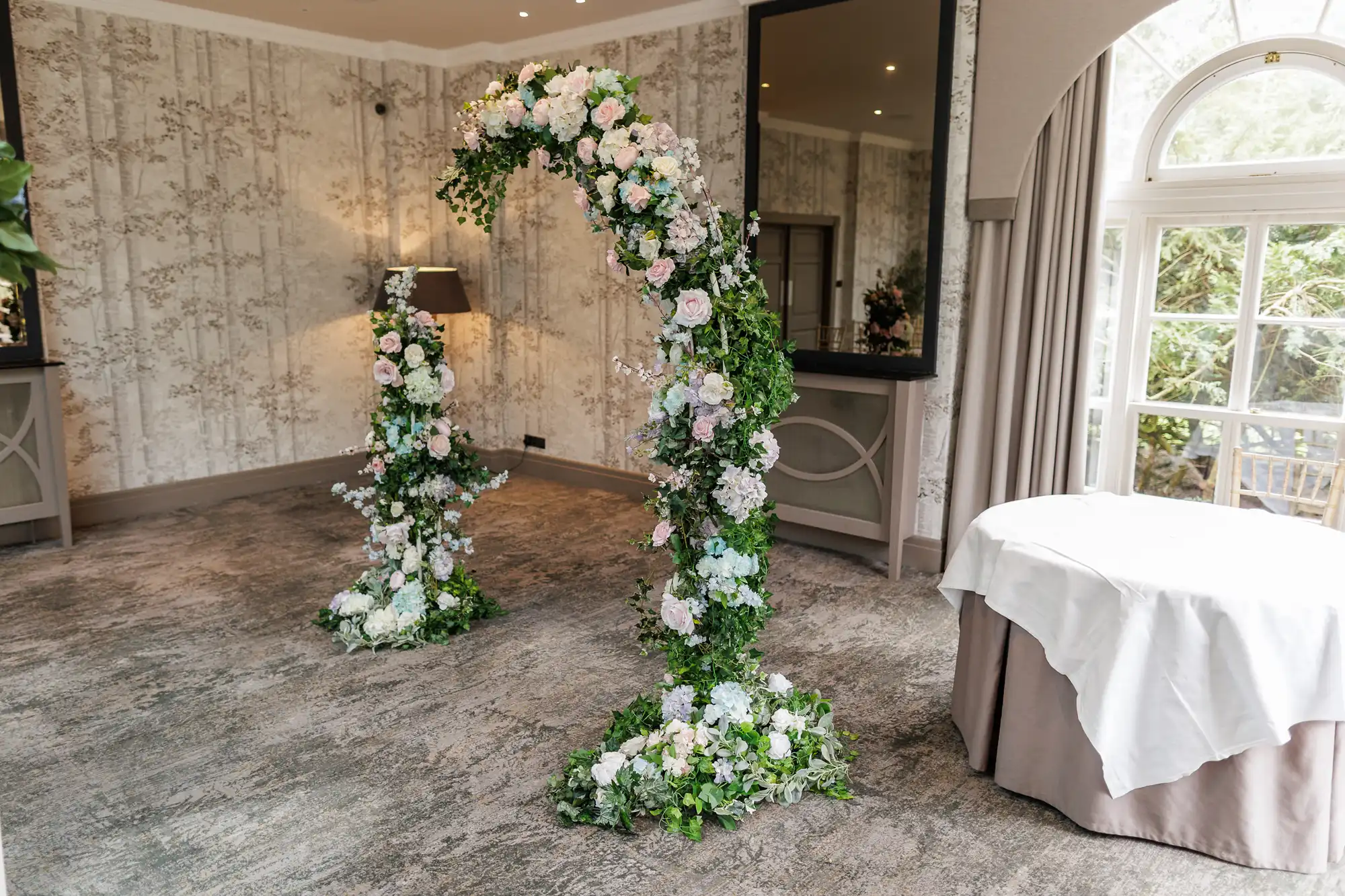 A floral arch and pillar decorated with white, pink, and blue flowers and lush greenery stand in a carpeted room with cream-patterned wallpaper. A round table with a white tablecloth is near a large arched window letting in natural light.