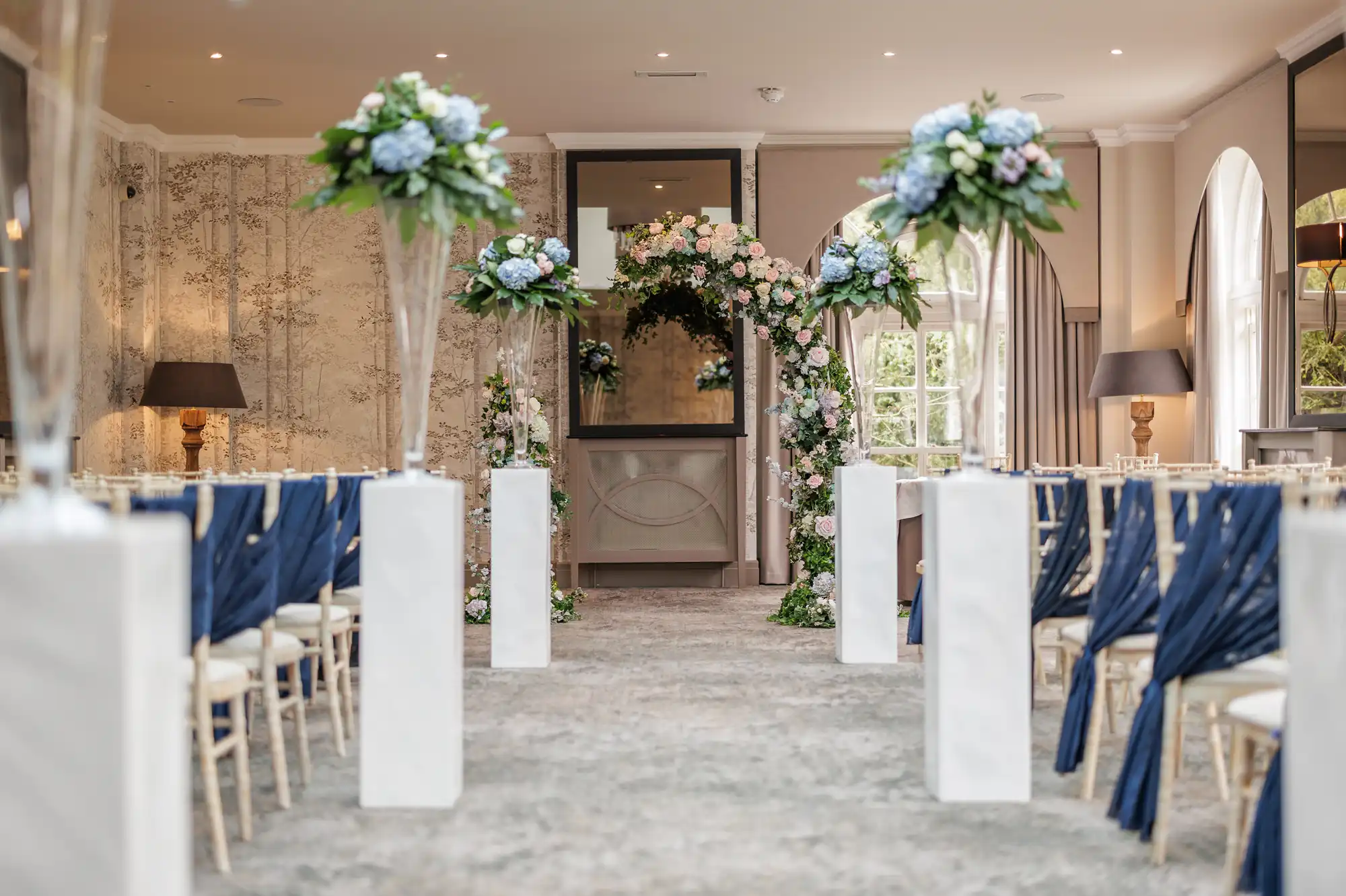 A wedding ceremony set-up with chairs draped in navy blue sashes, tall white stands topped with floral arrangements, and a flower-adorned arch at the front. Soft natural light enters through arched windows, creating an elegant and romantic atmosphere.