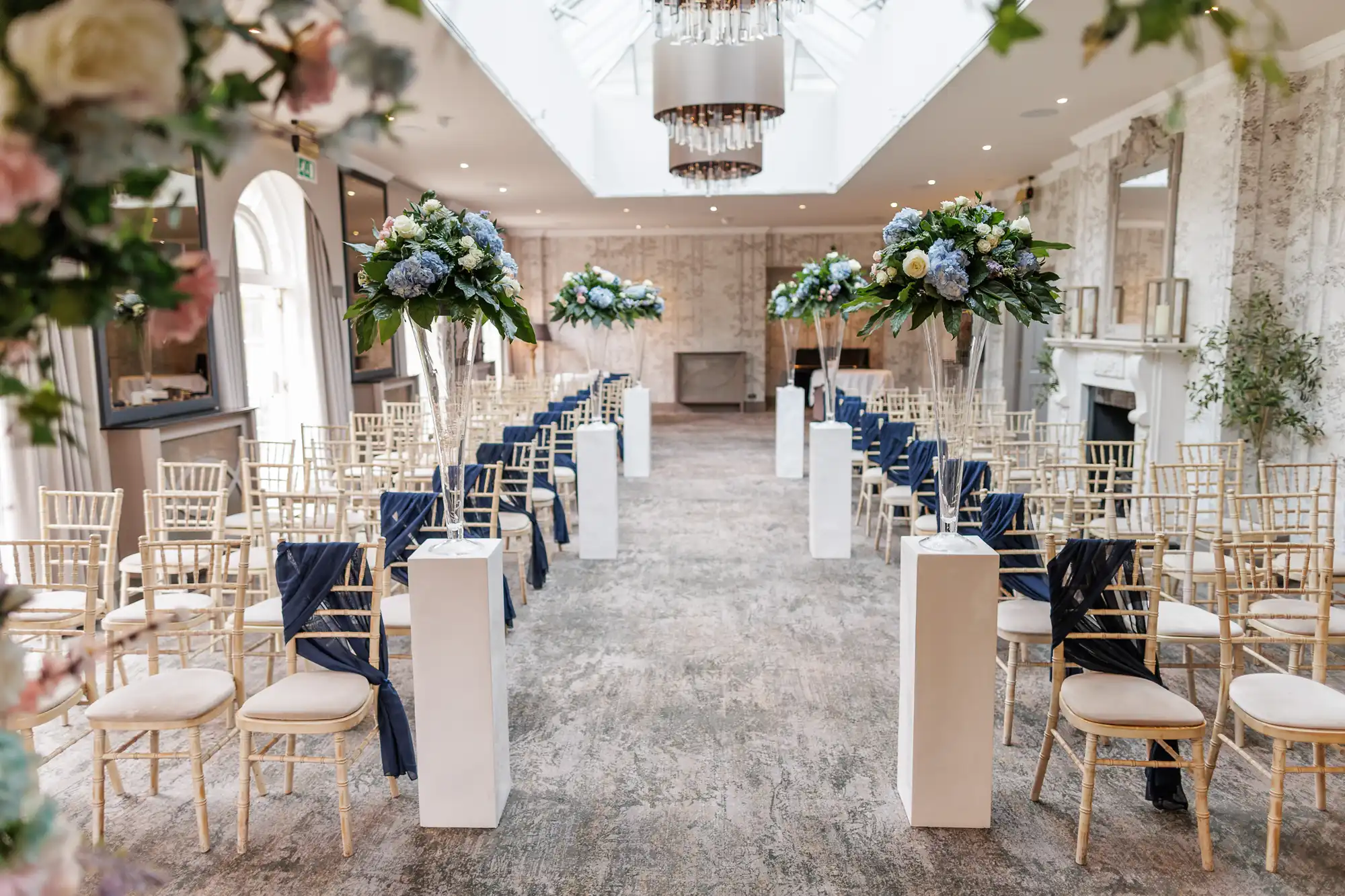 Elegant indoor wedding ceremony setup with rows of light wooden chairs, some draped with navy blue fabric. Tall floral arrangements with white and blue flowers line the aisle, which is carpeted. Large windows and chandeliers provide bright, natural light.