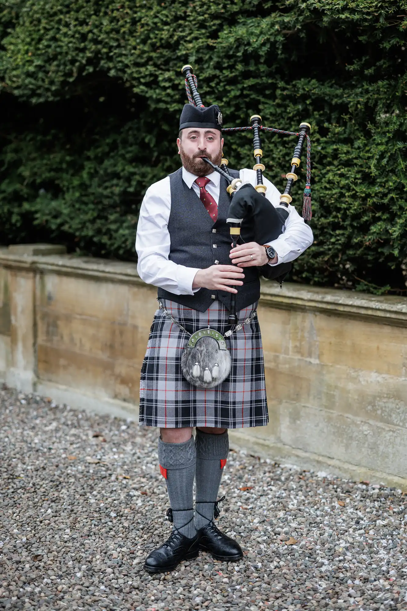 A man in traditional Scottish attire plays bagpipes outdoors. He wears a kilt, sporran, knee-high socks with flashes, a waistcoat, white shirt, tie, and a Glengarry hat. Behind him is a stone wall and green hedges.