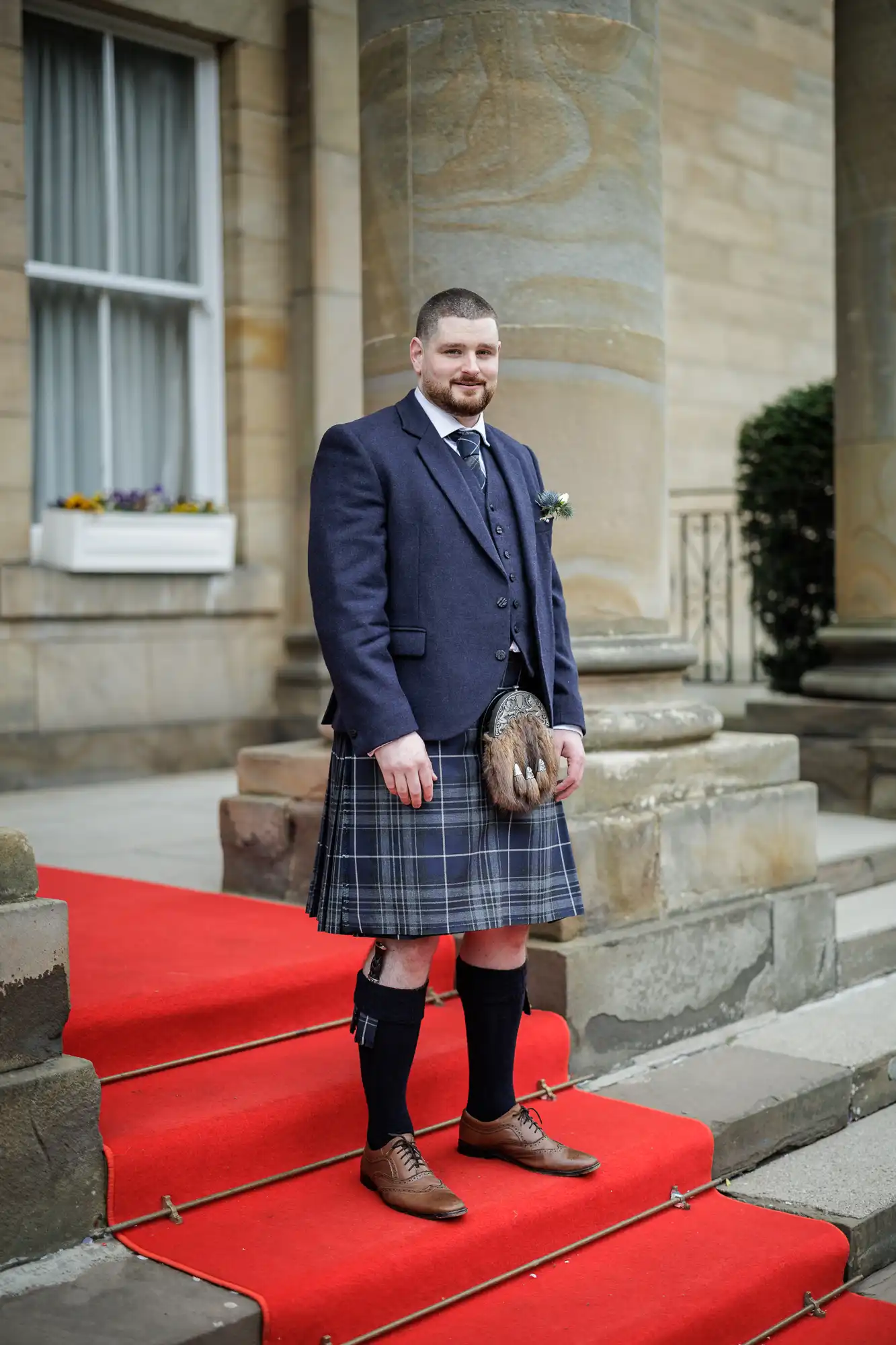 A man with short hair and a beard stands on a red carpet in front of a stone building. He wears a blue jacket, checked kilt, sporran, knee-high socks with garters, brown shoes, and a tie, with a buttonhole on his lapel.
