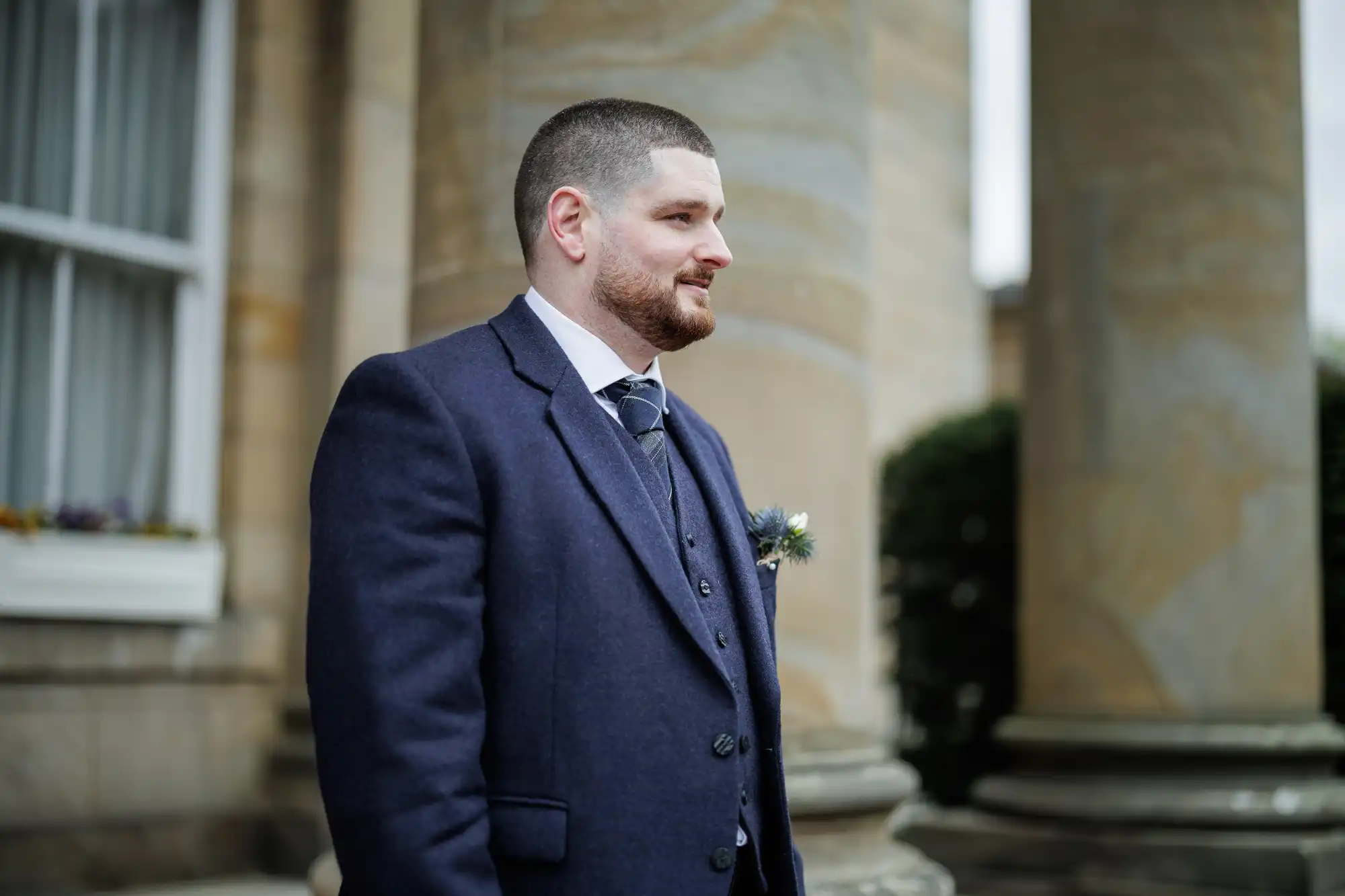 A man with short hair and a beard, wearing a navy suit with a tie and buttonhole, stands outdoors in front of large stone columns and a building with a window. He faces to the left and appears to be at a formal event.
