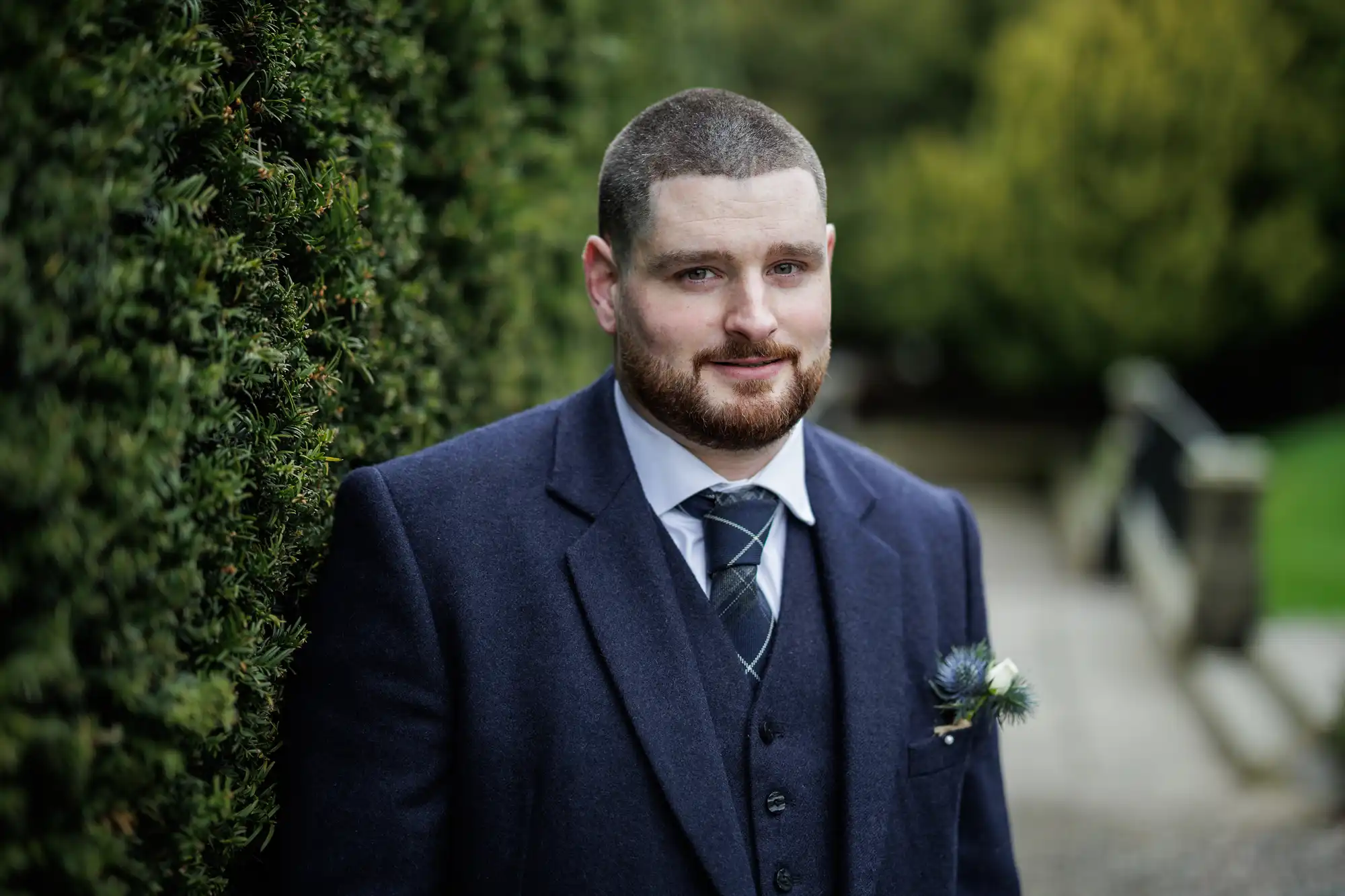 A man with short brown hair and beard, wearing a dark blue suit, white shirt, and checked tie, stands outdoors by a hedge. He is slightly smiling, with a buttonhole pinned to his lapel. The background is blurred greenery and a stone path.