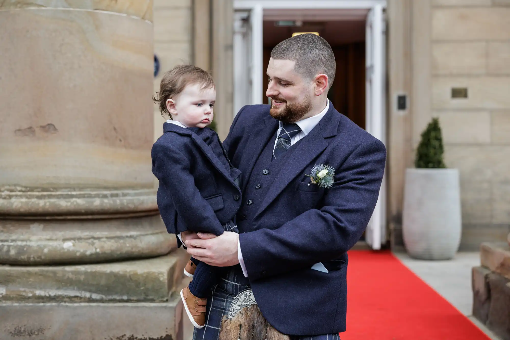 A bearded man in a navy suit and kilt smiles whilst holding a young boy, also in a matching navy suit, outside a stone building with a large column and a red carpet leading to an open door in the background.