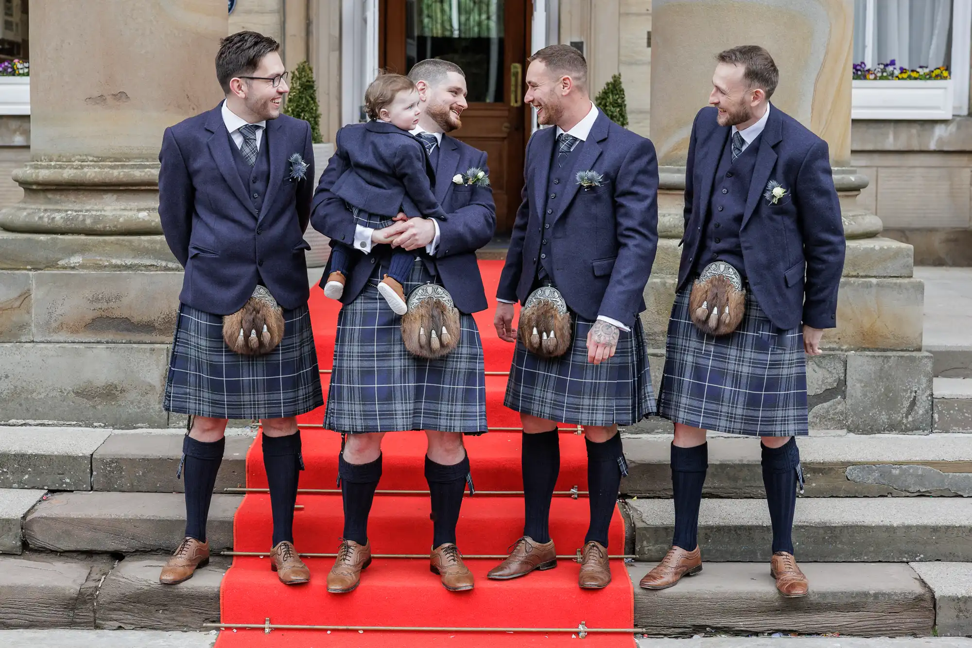 Four men in matching blue jackets, plaid kilts, and sporrans stand on a red carpet outside a stone building. One man holds a small child dressed similarly. All are smiling and appear to be enjoying a celebratory moment together.