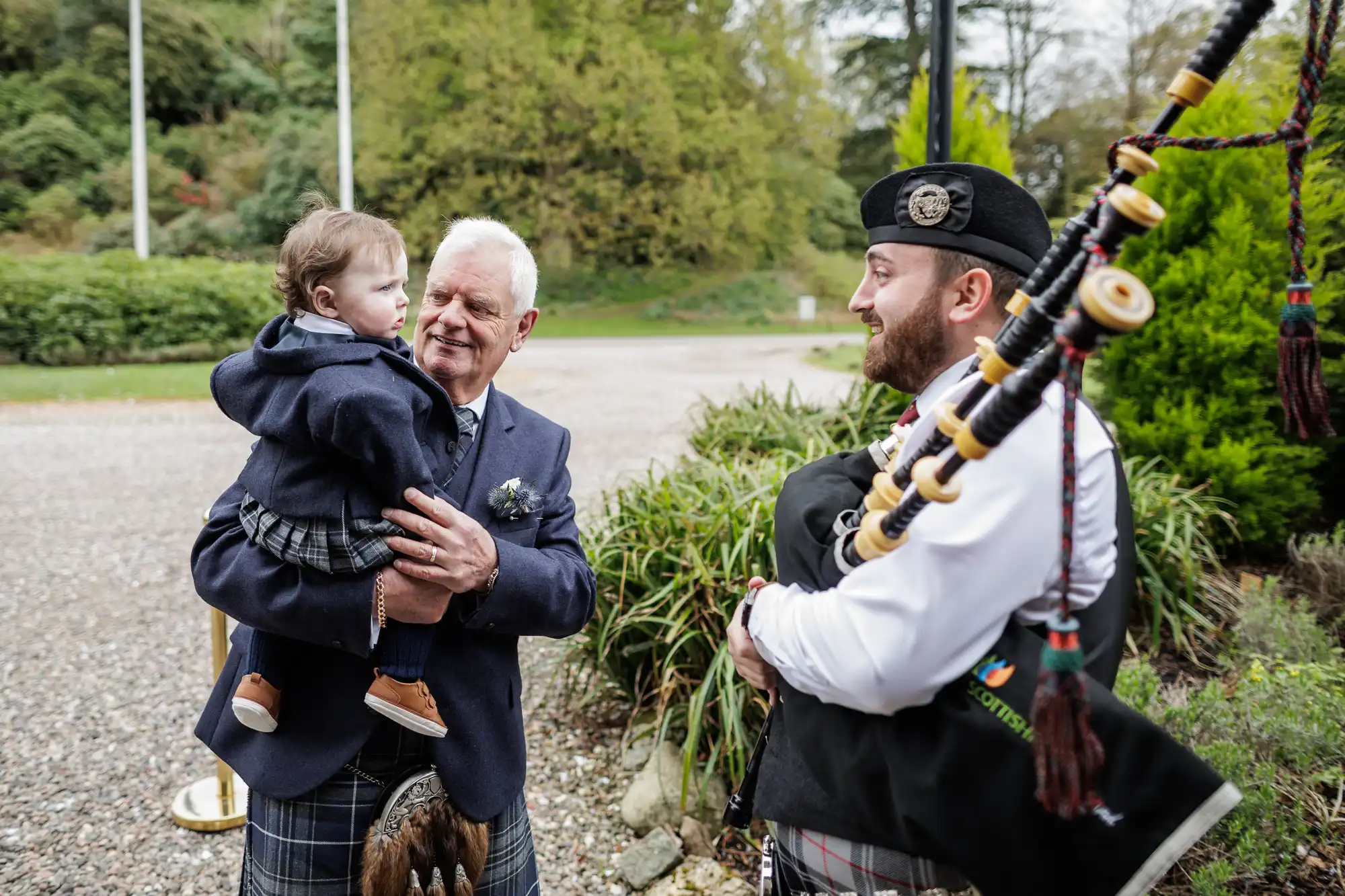An older man in a kilt holds a young child while smiling outdoors. A bagpiper wearing a traditional uniform stands nearby, holding bagpipes. Trees and greenery form the background, creating a cheerful and festive atmosphere.