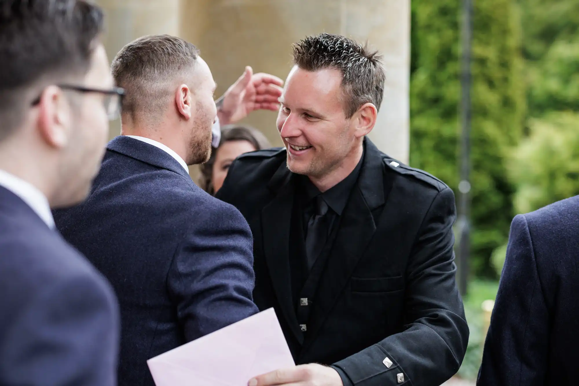 Two men in suits greet each other warmly outdoors, with one man smiling and extending his arm for a hug. Another man in glasses is partially visible on the left. Lush greenery and a stone pillar are in the background.