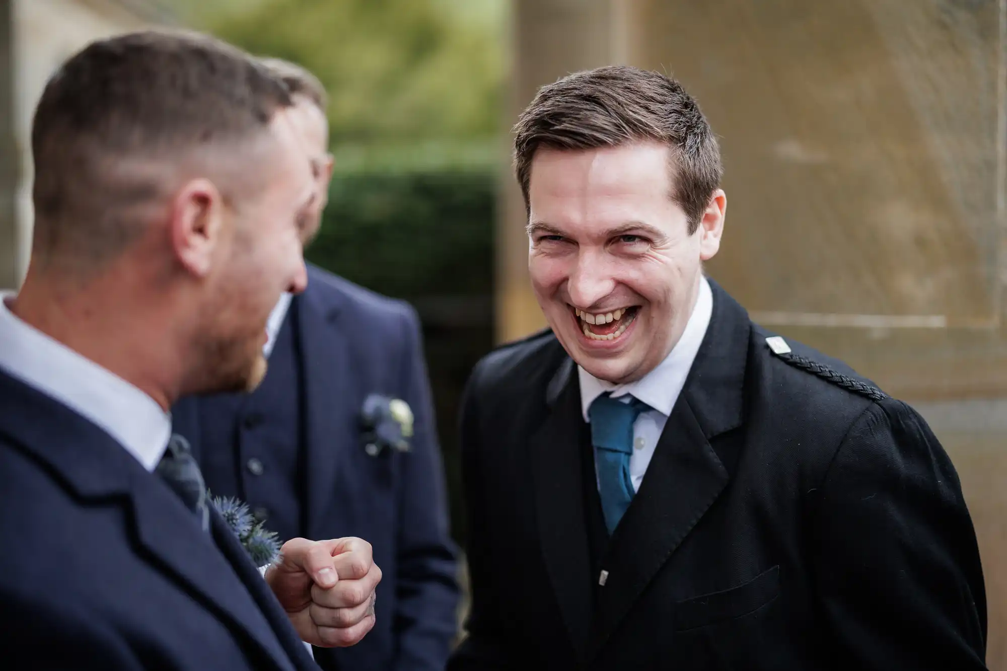 Two men in suits are laughing and talking outdoors. The man on the right, with short dark hair, is grinning widely, while the man on the left faces him and smiles. Another person in a suit is blurred in the background. The setting appears to be formal or celebratory.