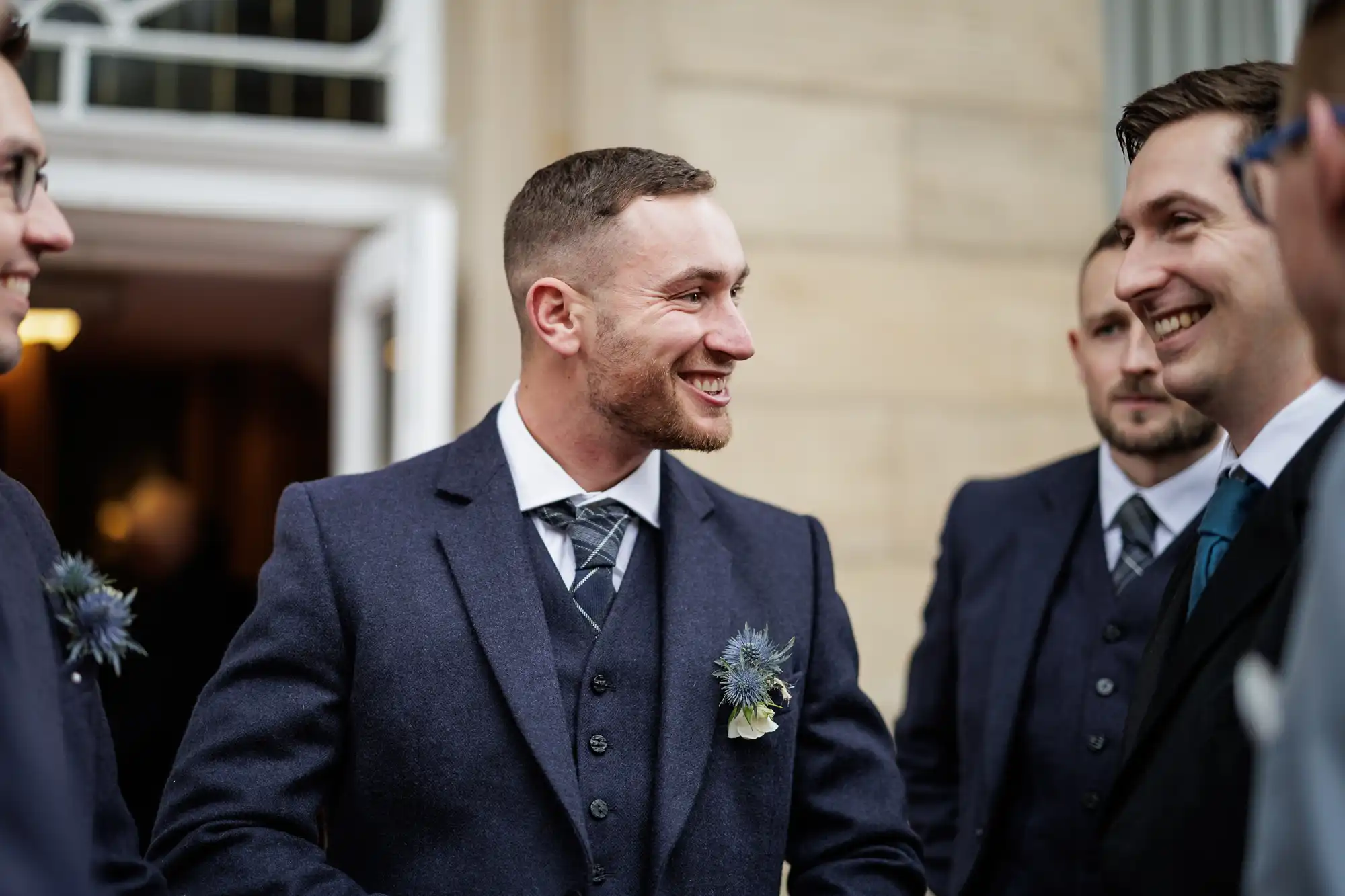 A smiling man in a navy suit with a buttonhole stands outside talking to other men in suits. They appear to be at a formal event, possibly a wedding, and are gathered near a building with beige stone walls and large windows.