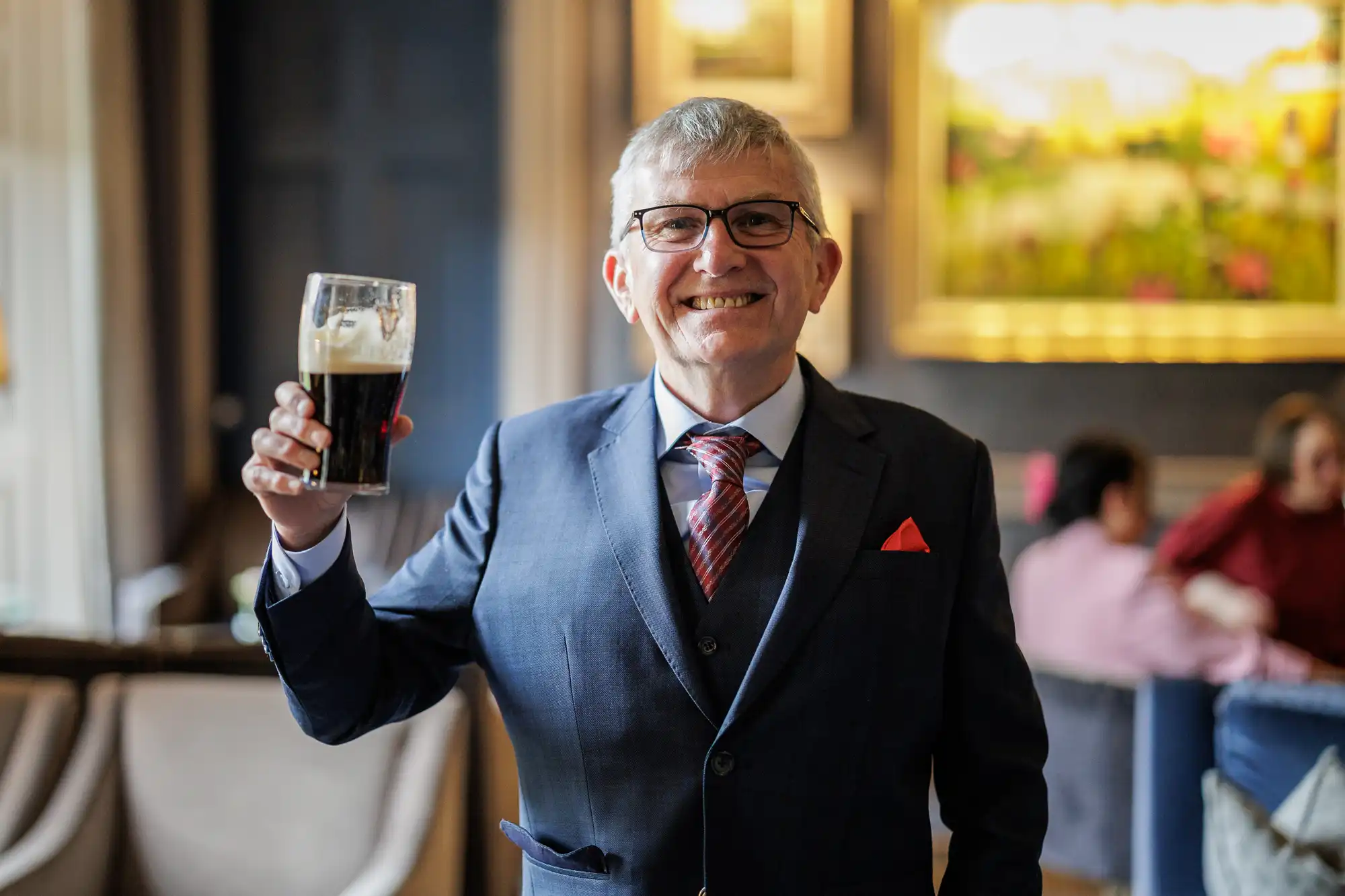 An older man in a blue suit, red tie, and glasses smiles whilst holding up a pint of dark beer. He stands indoors in a warmly lit room with framed artwork and people seated in the blurred background.