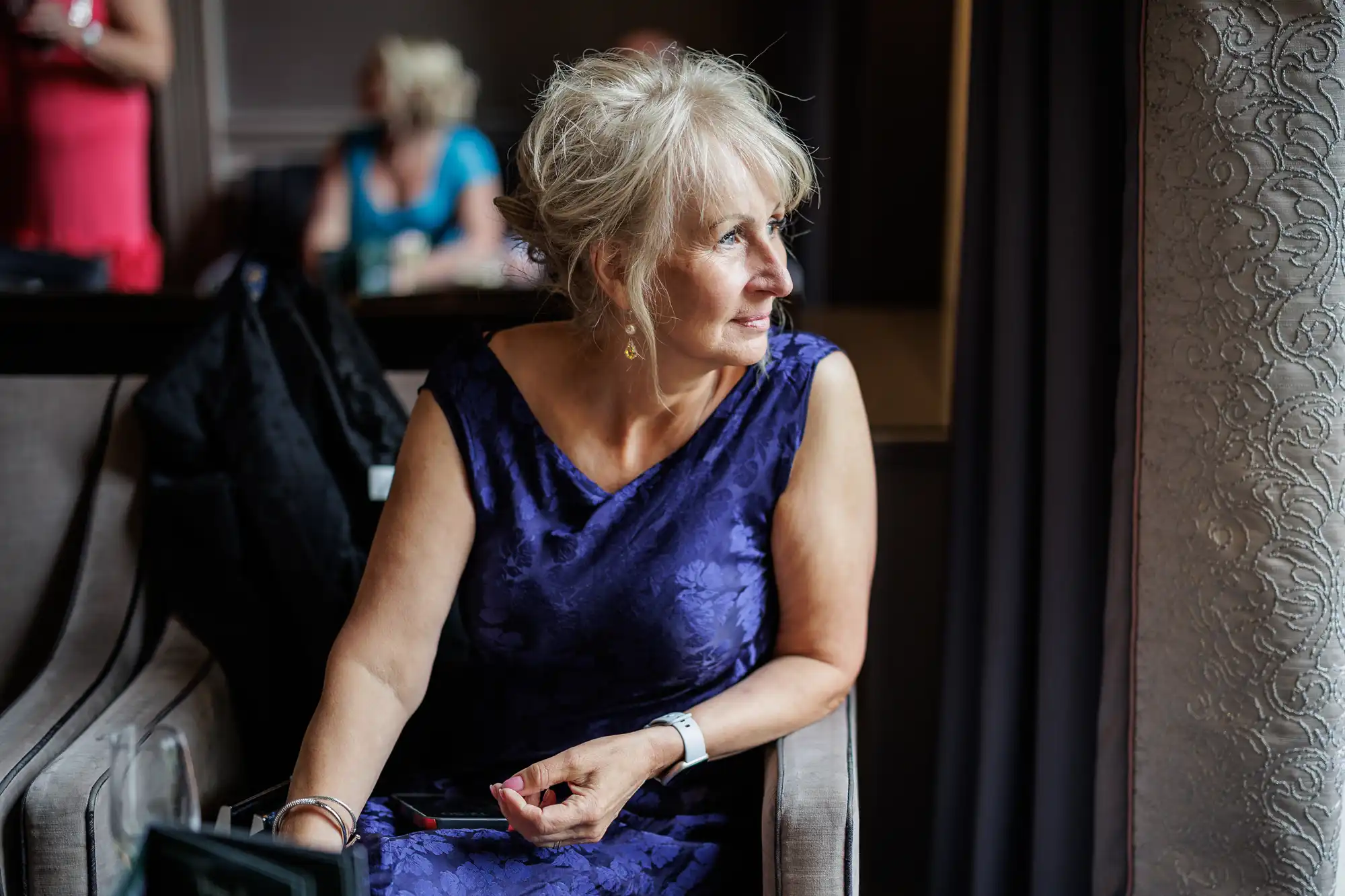 A woman with blonde hair in an updo, wearing a sleeveless purple dress, sits on a chair and looks thoughtfully out of a window. Soft light falls on her face. Other people are blurred in the background. Elegant decor and curtains are visible.