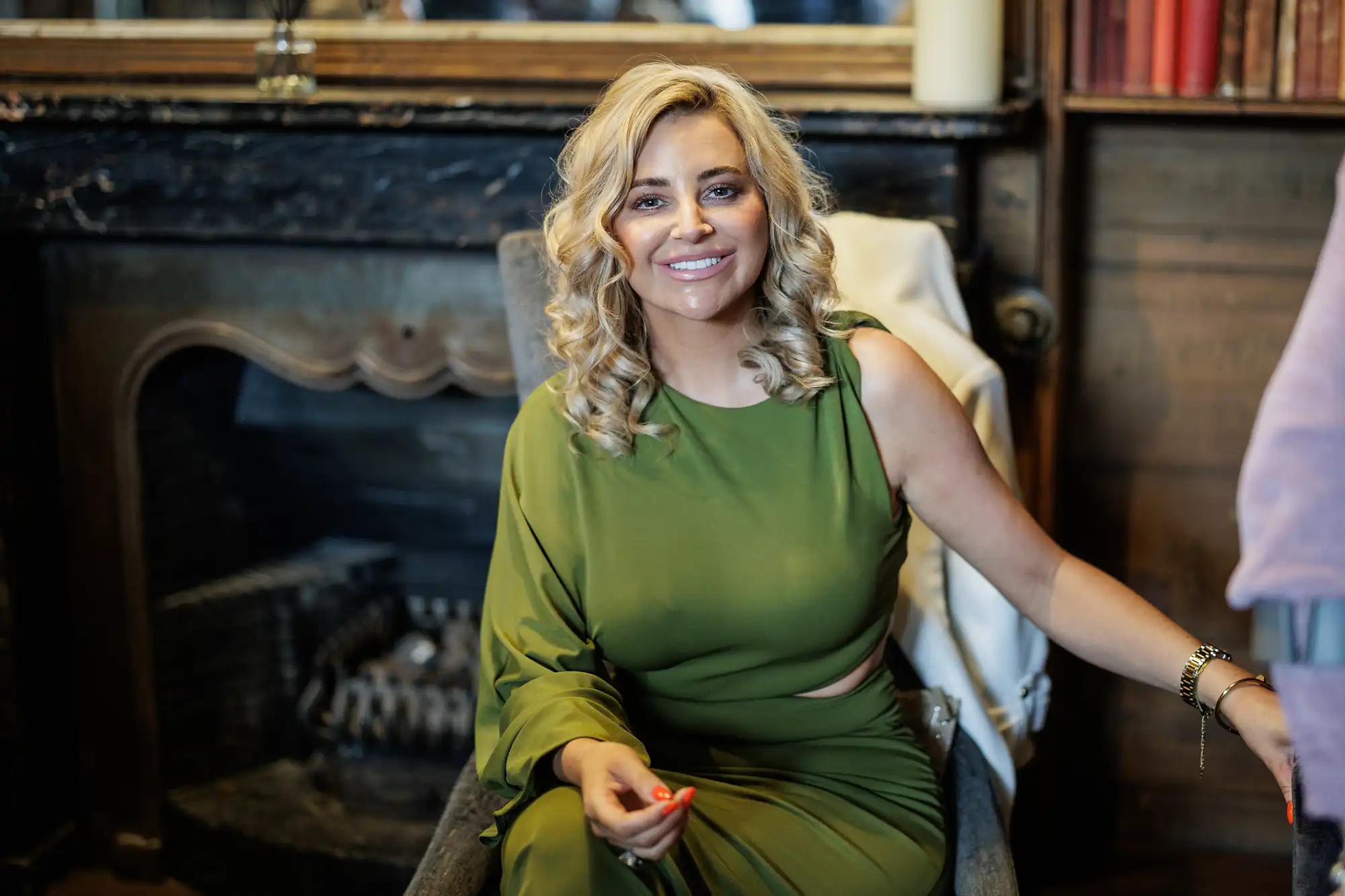 A woman with curly blonde hair wearing a sleeveless green dress sits smiling in a chair indoors. A fireplace with a black mantel and wooden furnishings are in the background. She rests one arm on the chair and has orange nail varnish on her visible hand.