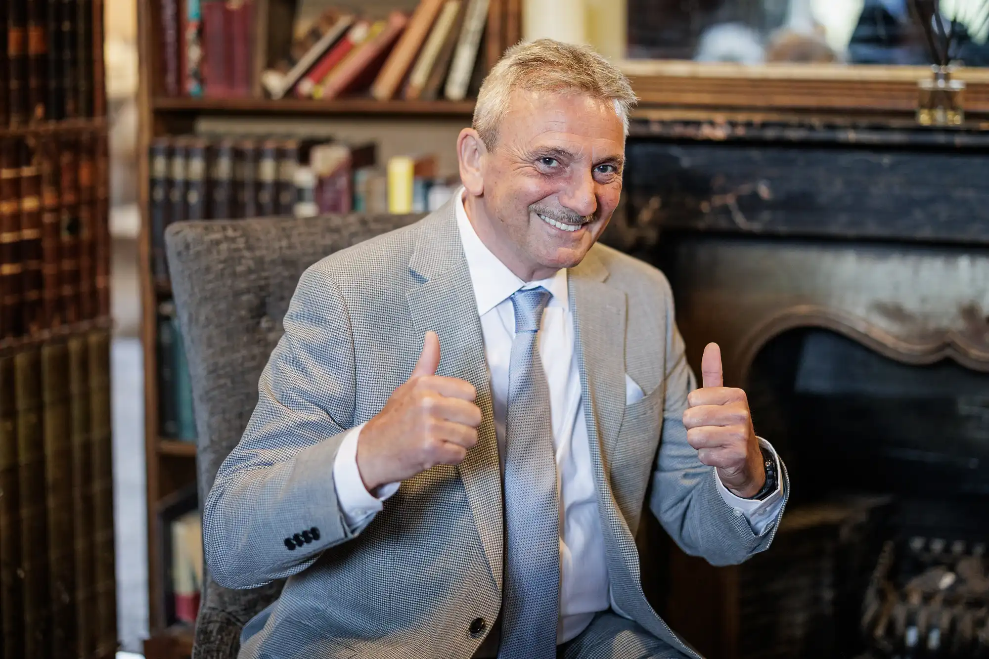 An older man in a light grey suit, white shirt, and tie sits on a chair indoors, smiling and giving two thumbs up. Behind him are bookshelves filled with books and a dark fireplace, creating a cosy, warm setting.