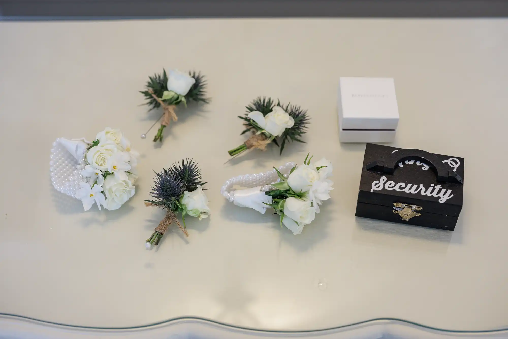 Five buttonholes with white roses and greenery are arranged on a white surface. Nearby are a pearl bracelet corsage, a small white box, and a black ring bearer case labelled “Ring Security” with a gold lock.