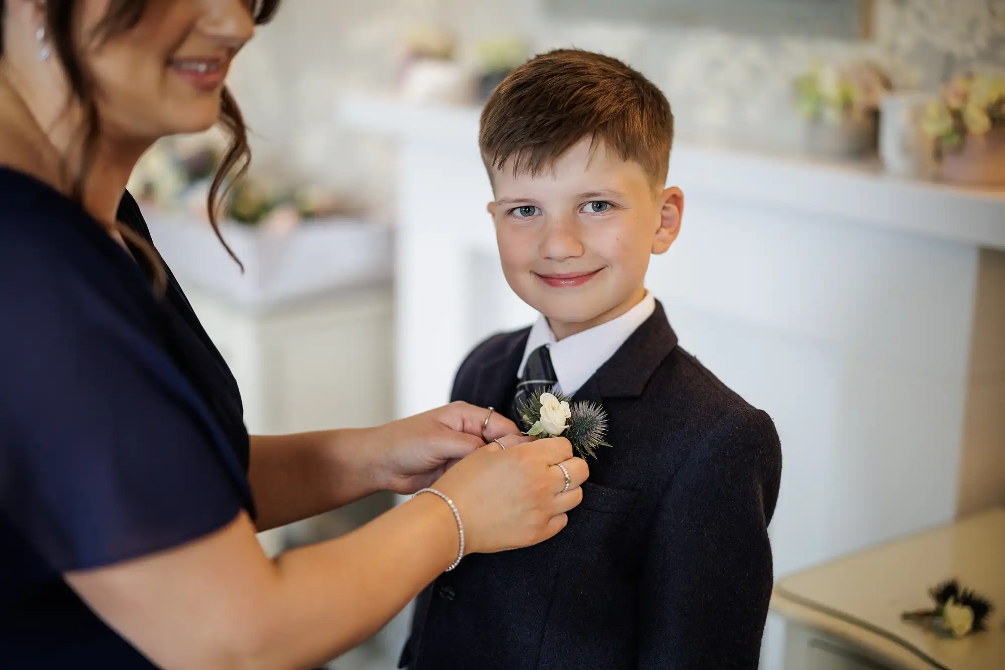 A woman in a navy dress pins a buttonhole to a young boy’s dark suit jacket. The boy, with short brown hair, smiles at the camera. The background is softly blurred, with pastel flowers and a white fireplace visible.