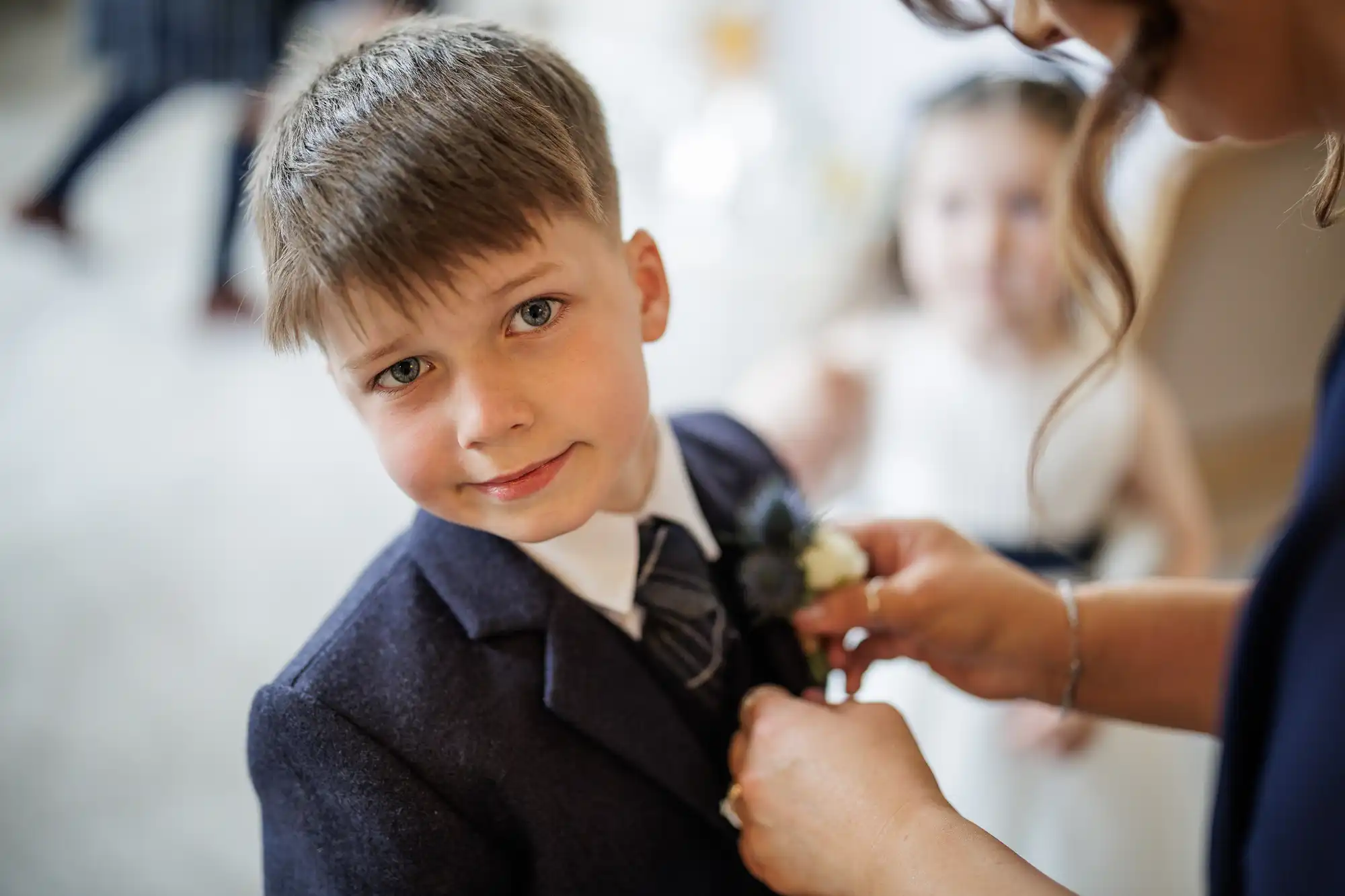 A young boy in a dark suit and tie looks at the camera as an adult adjusts a buttonhole on his lapel. In the blurred background, a girl in a white dress is visible. The setting appears formal, possibly a wedding or special occasion.