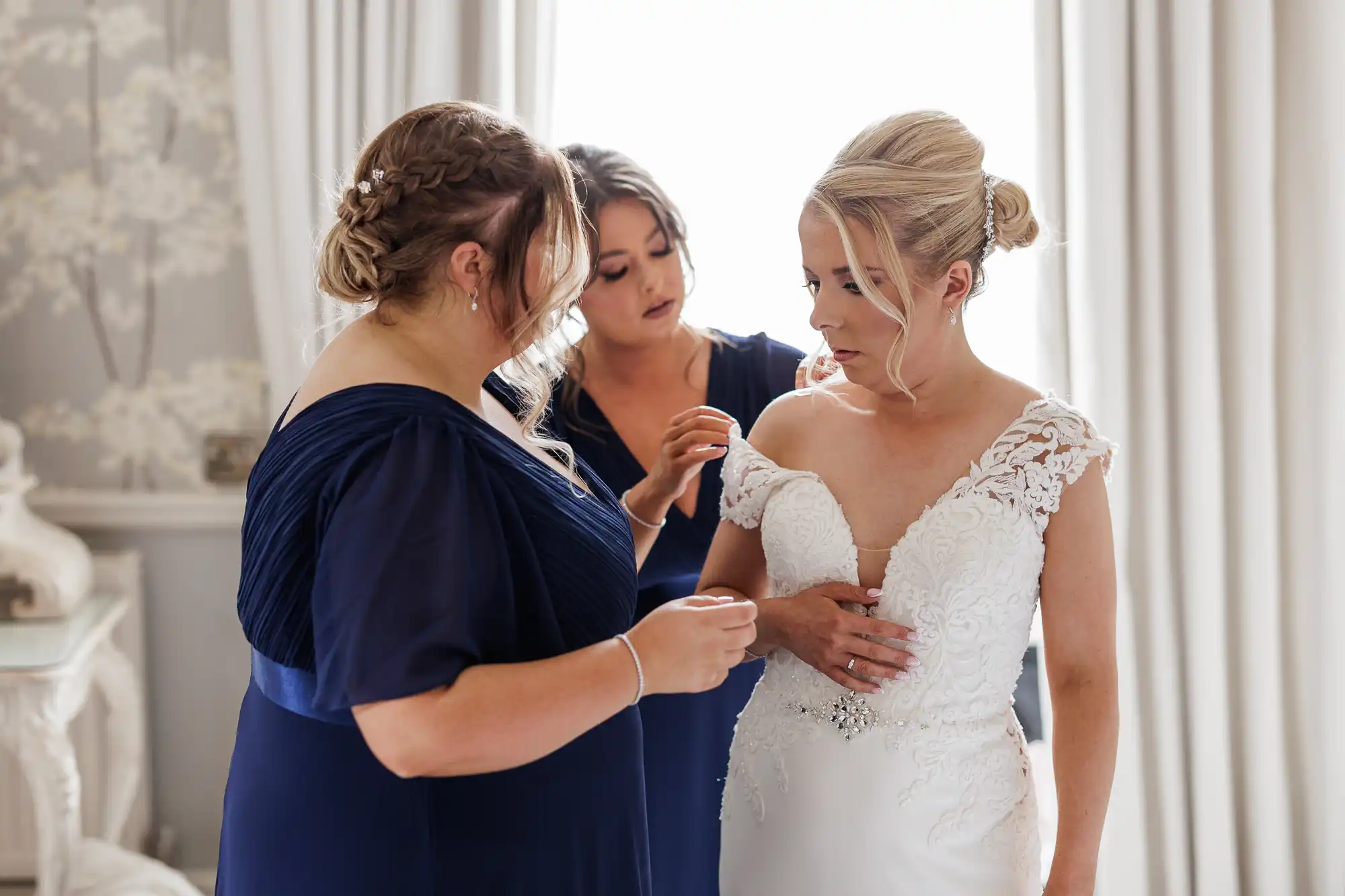 Three women stand in a softly lit room. The woman on the right wears a white lace wedding dress, looking down thoughtfully. The two women beside her in navy dresses help adjust her dress, preparing her for the wedding. Cream curtains and elegant décor are in the background.