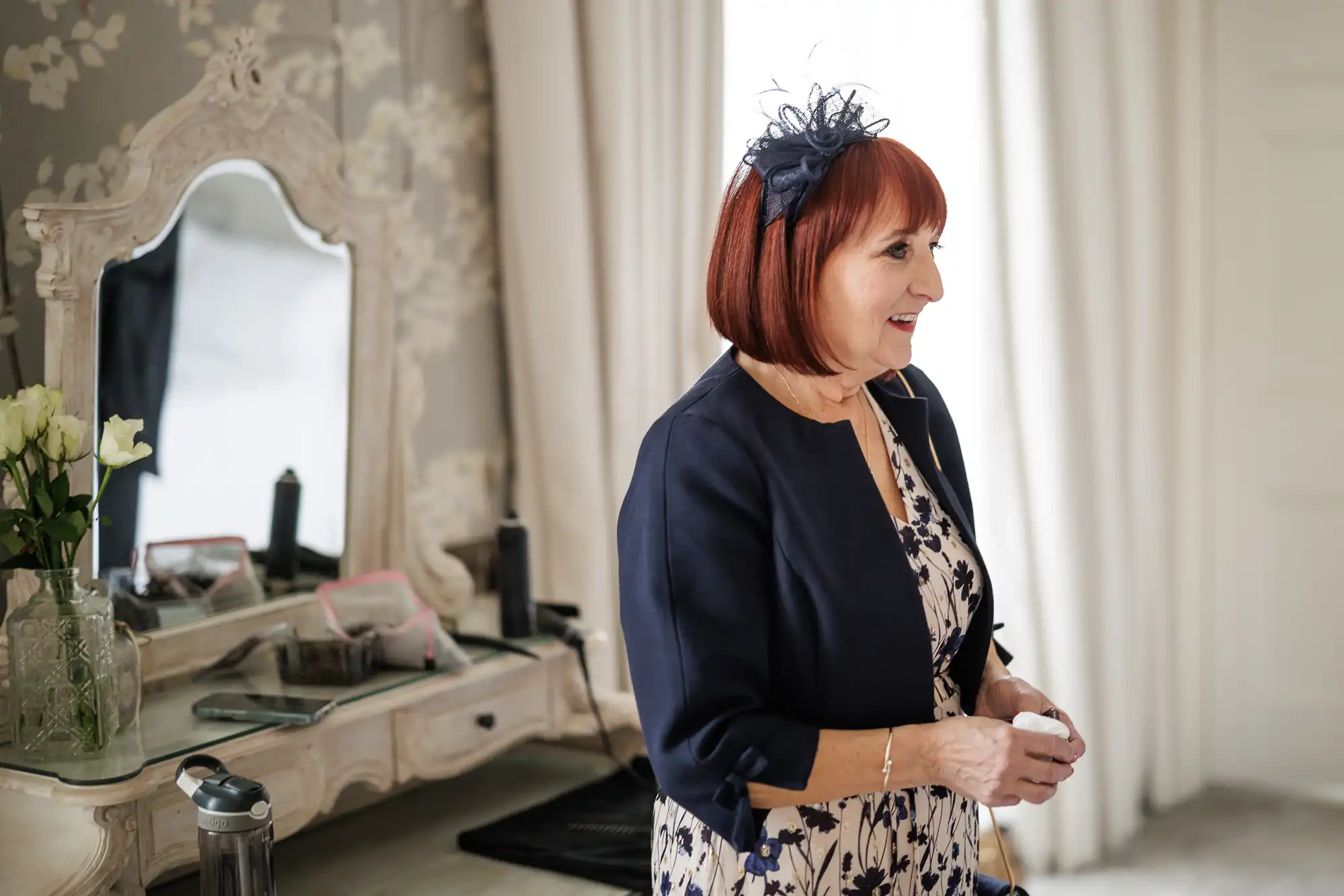 An older woman with red hair, wearing a navy fascinator, navy jacket, and patterned dress, stands smiling indoors. Behind her is a vintage-style dressing table with a mirror, white flowers, and various items, with cream curtains in the background.