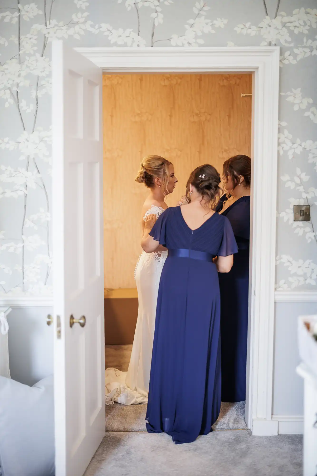 A bride in a white dress stands in a doorway, facing two women in matching navy blue gowns, who are helping her get ready. The scene is set in a softly lit room with floral wallpaper and a partially open white door.
