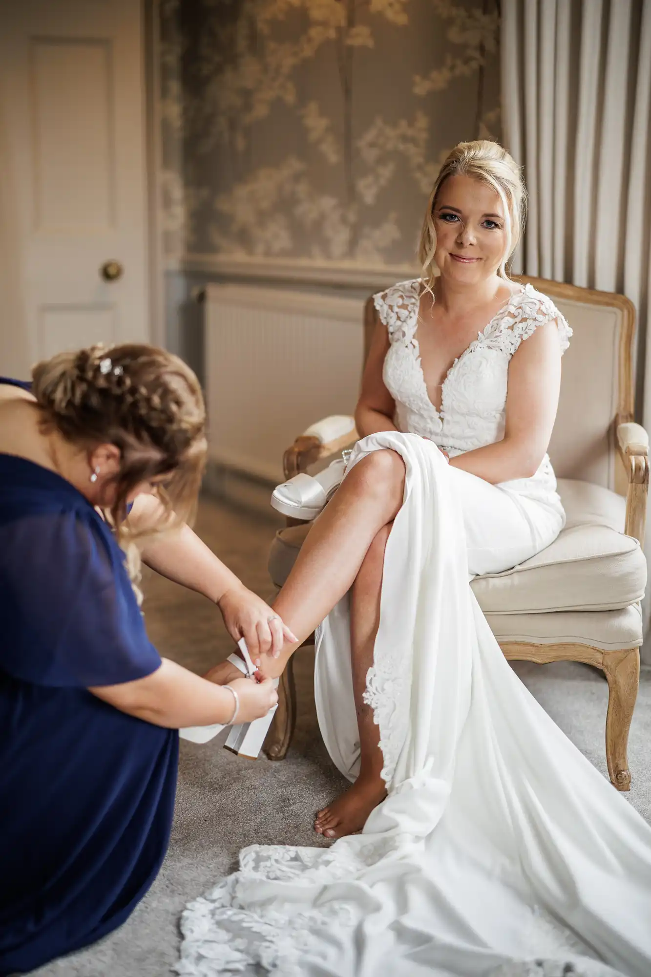 A bride in a white lace wedding dress sits on a cream chair, smiling softly. Another woman in a navy dress kneels, helping her put on white shoes. The room has neutral decor, floral wallpaper, and soft lighting from a window with curtains.