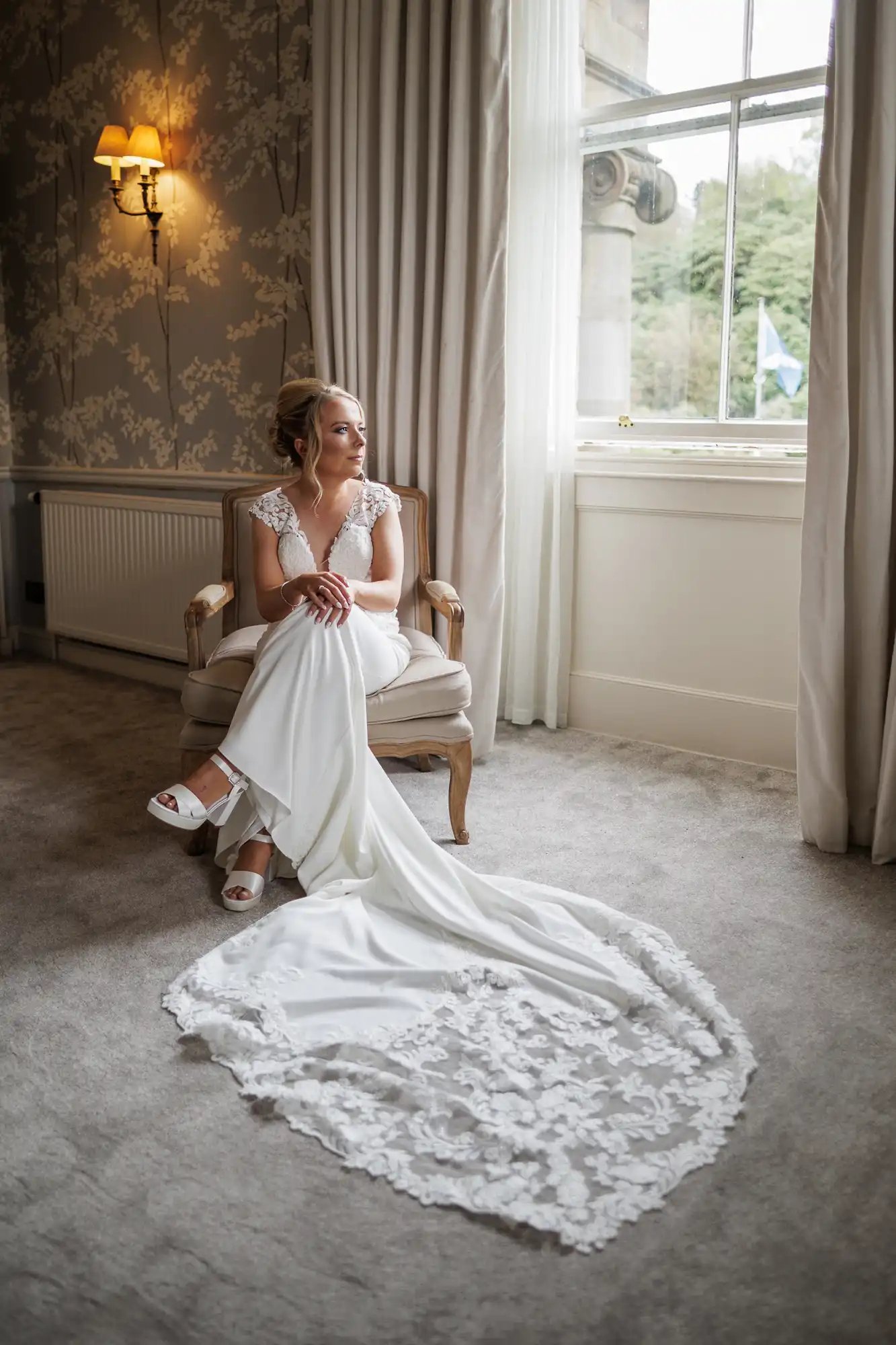 A bride in a white gown with lace details sits on a cushioned chair by a window with sheer curtains. She looks thoughtfully outside, hands clasped, with her dress elegantly spread out on the carpeted floor. A wall sconce glows behind her.