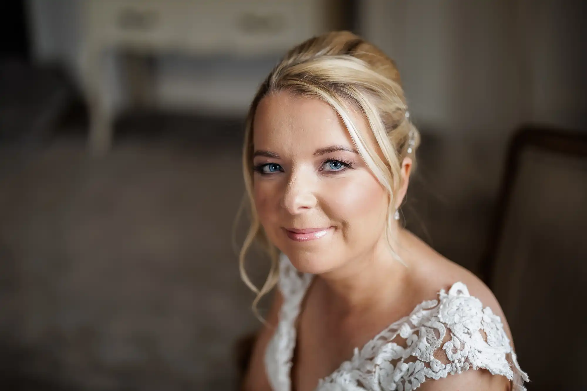 A woman with blonde hair in an elegant updo, wearing a white lace wedding dress, smiles softly at the camera. The background is softly blurred, highlighting her make-up and bright blue eyes, giving a warm and serene atmosphere.