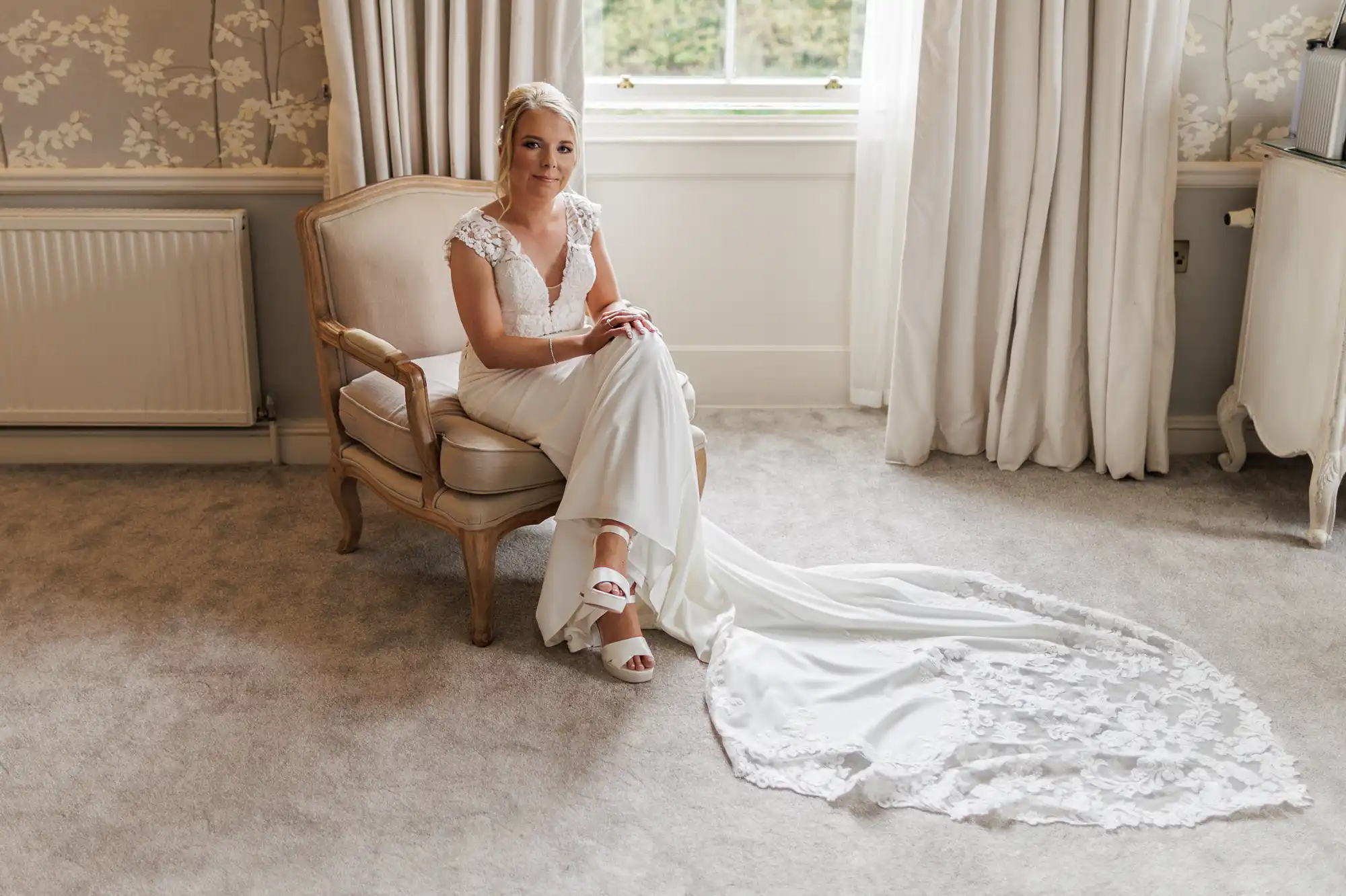 A bride in a white lace wedding dress sits on an elegant beige armchair in a bright room with neutral décor, pale carpet, and floral wallpaper. She looks at the camera, hands folded, with her dress train elegantly spread out on the floor.