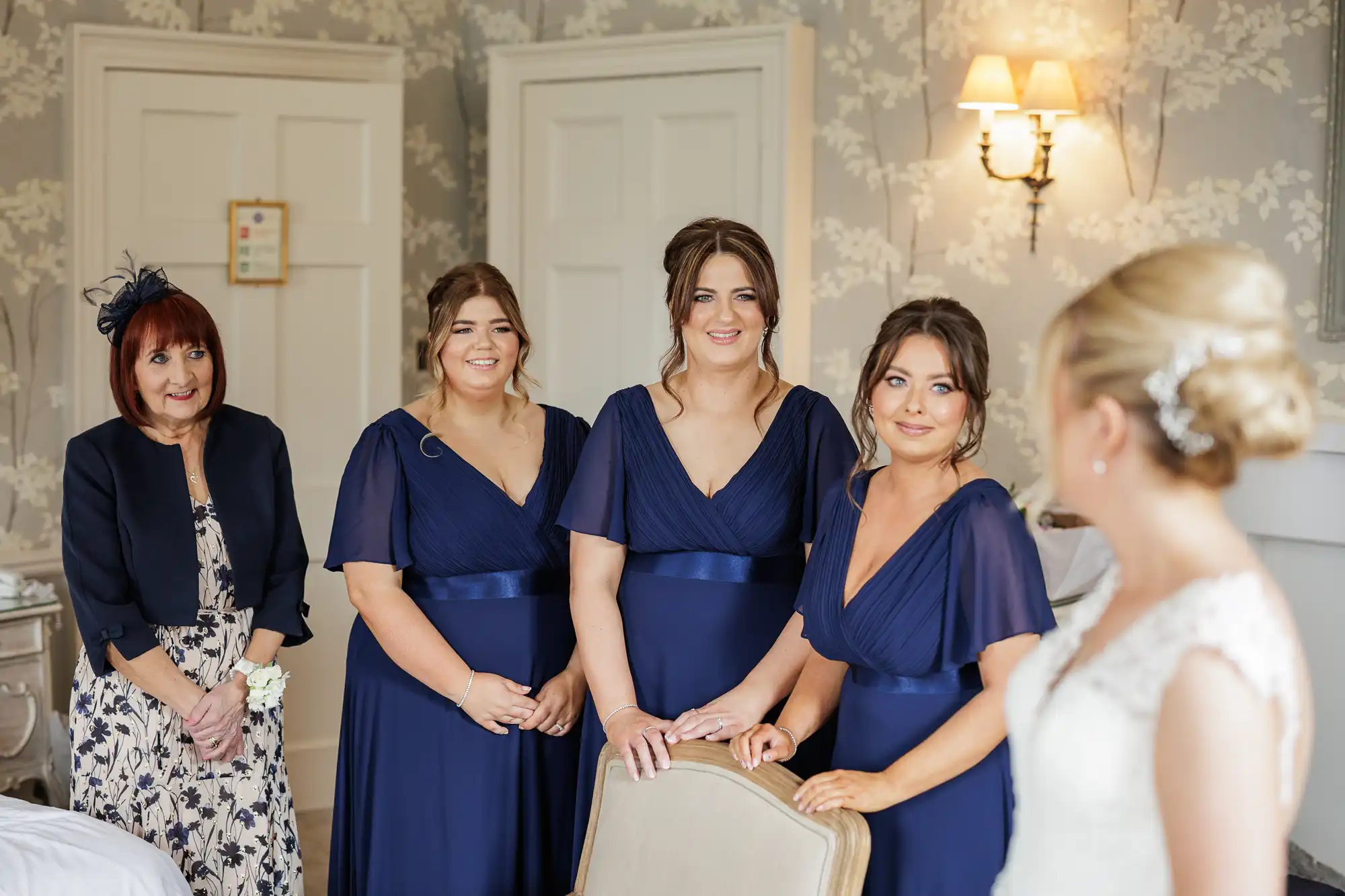 Four women in navy dresses and one woman in a floral dress and navy jacket stand in a softly lit room, smiling at a bride in a white gown with an ornate hairpiece, whose back is to the camera. The mood is joyful and anticipatory.