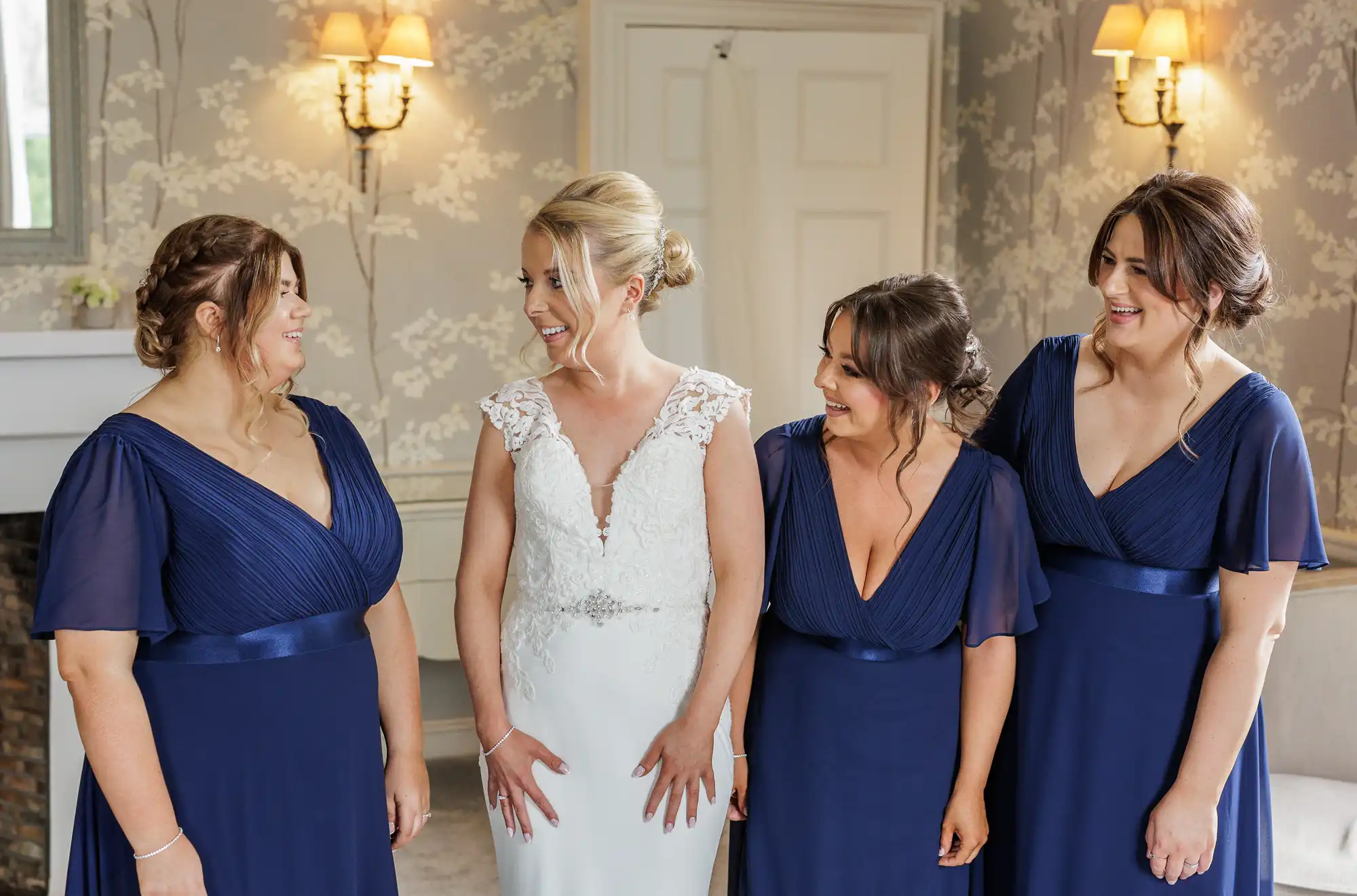 Four women stand indoors, smiling and chatting. The central woman wears a white bridal dress with lace details, while the three women around her wear matching navy blue bridesmaid dresses. The room has elegant wallpaper, wall lamps, and a white door in the background.