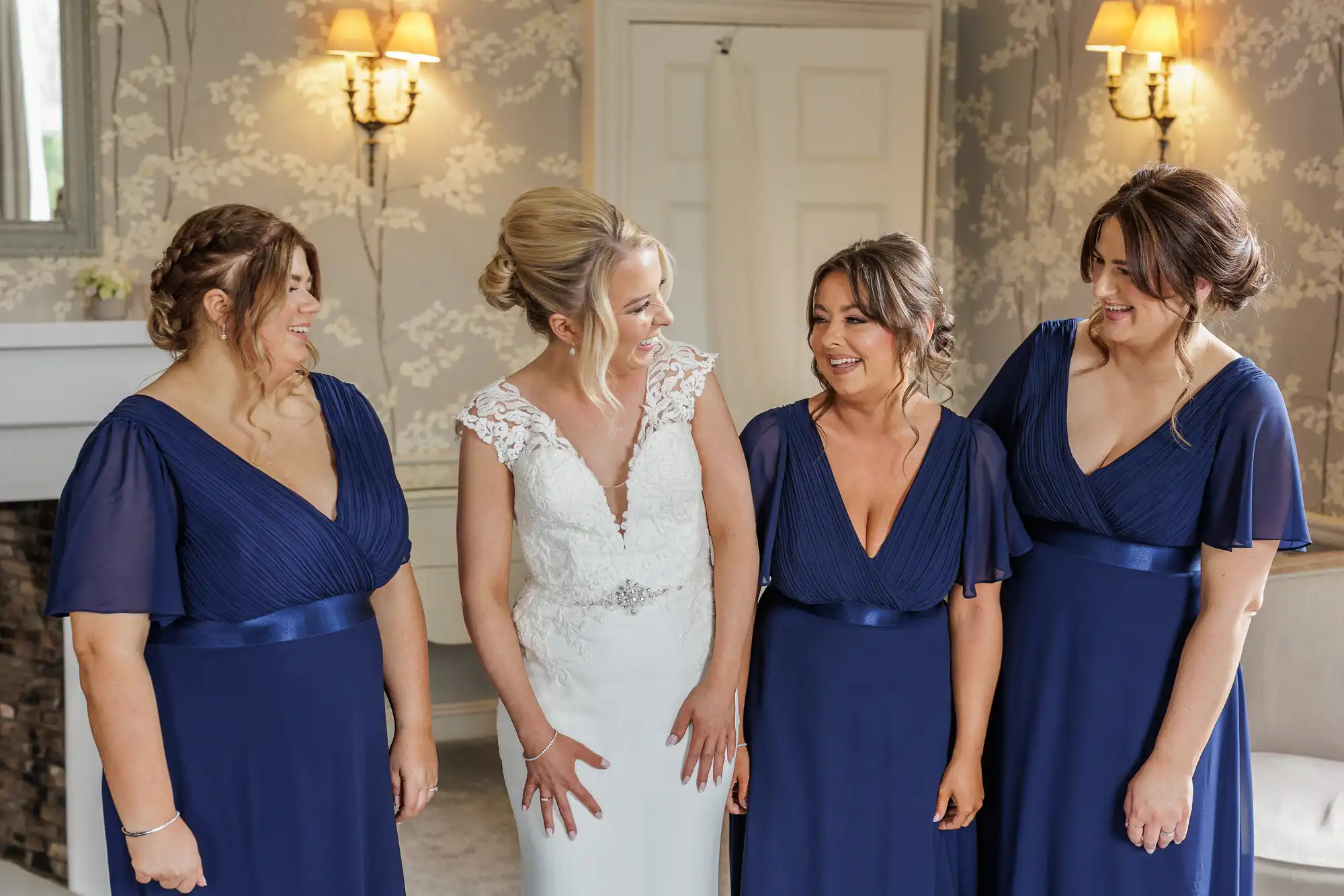 A bride in a white gown with lace details stands smiling with three bridesmaids in matching navy blue dresses. They are indoors, under warm wall lights, sharing a joyful moment together before the wedding.