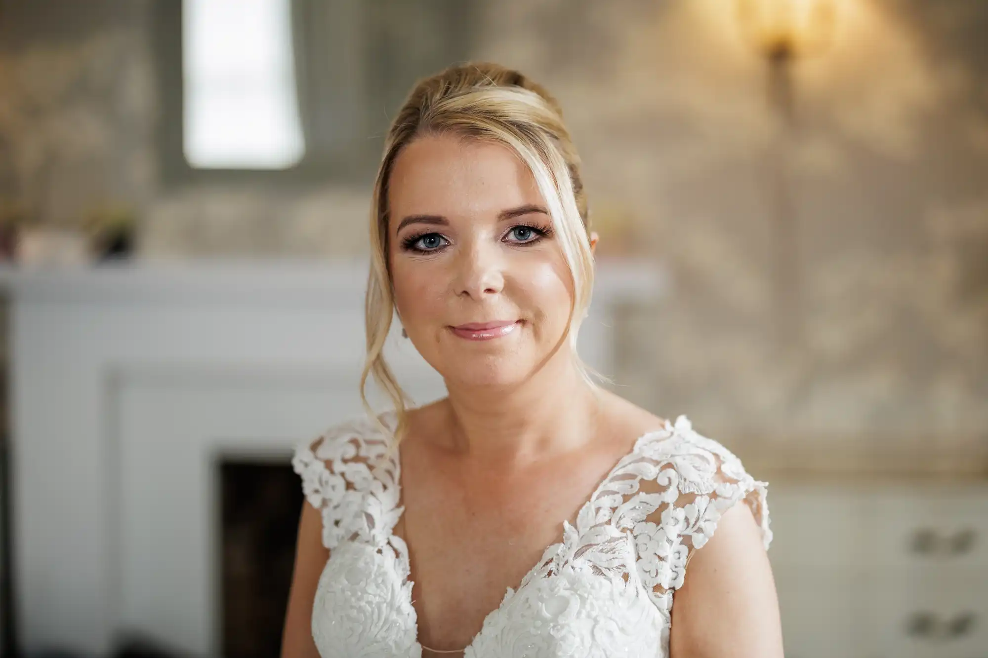 A woman with blonde hair in an updo, wearing a white lace wedding dress, smiles gently at the camera. The background is softly focused, showing a light-coloured room with a fireplace and warm lighting.