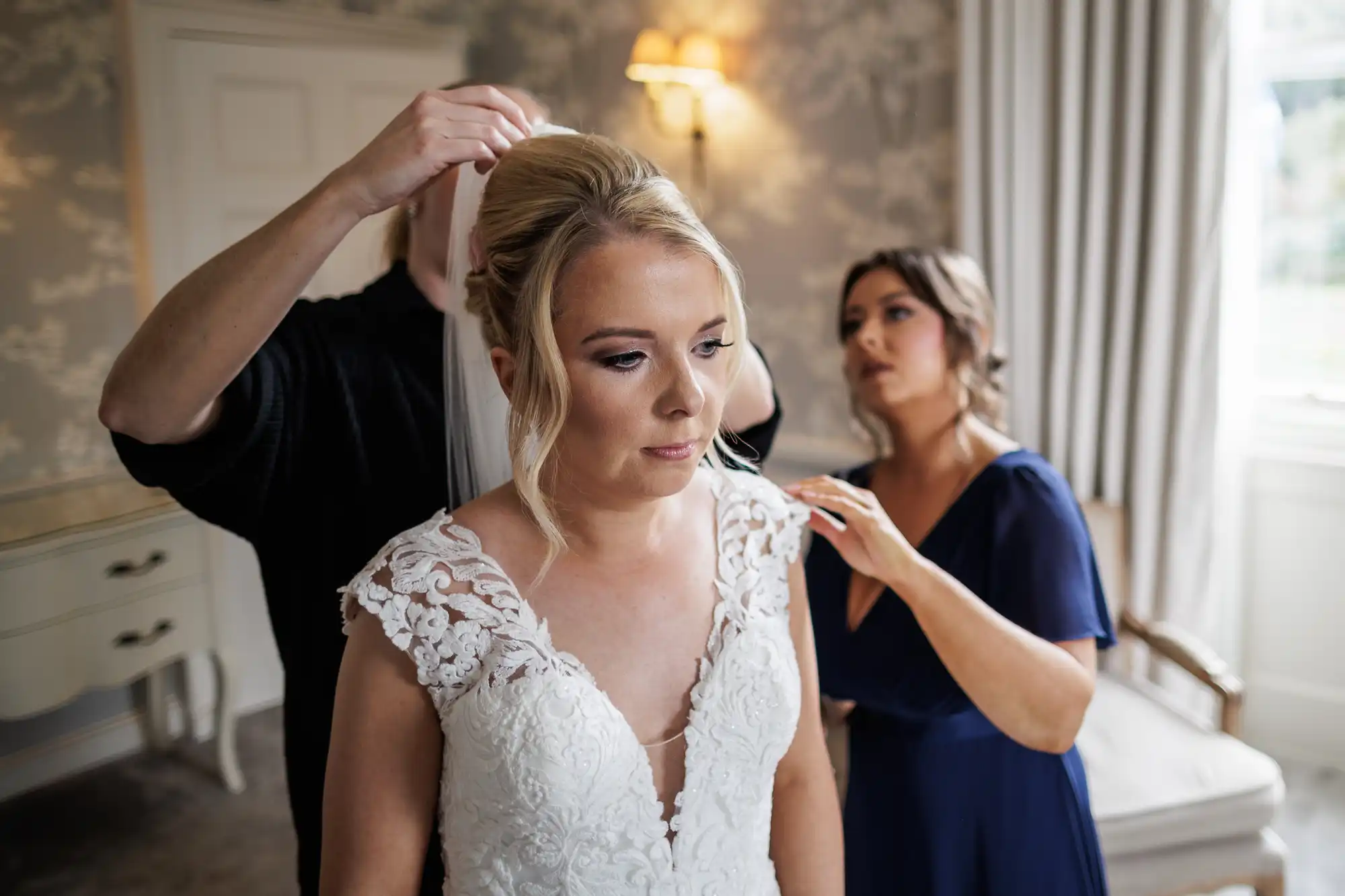A bride in a white lace wedding dress stands calmly as someone adjusts her veil and another woman in a navy dress helps with her gown. They are in a softly lit room with cream walls and large windows.