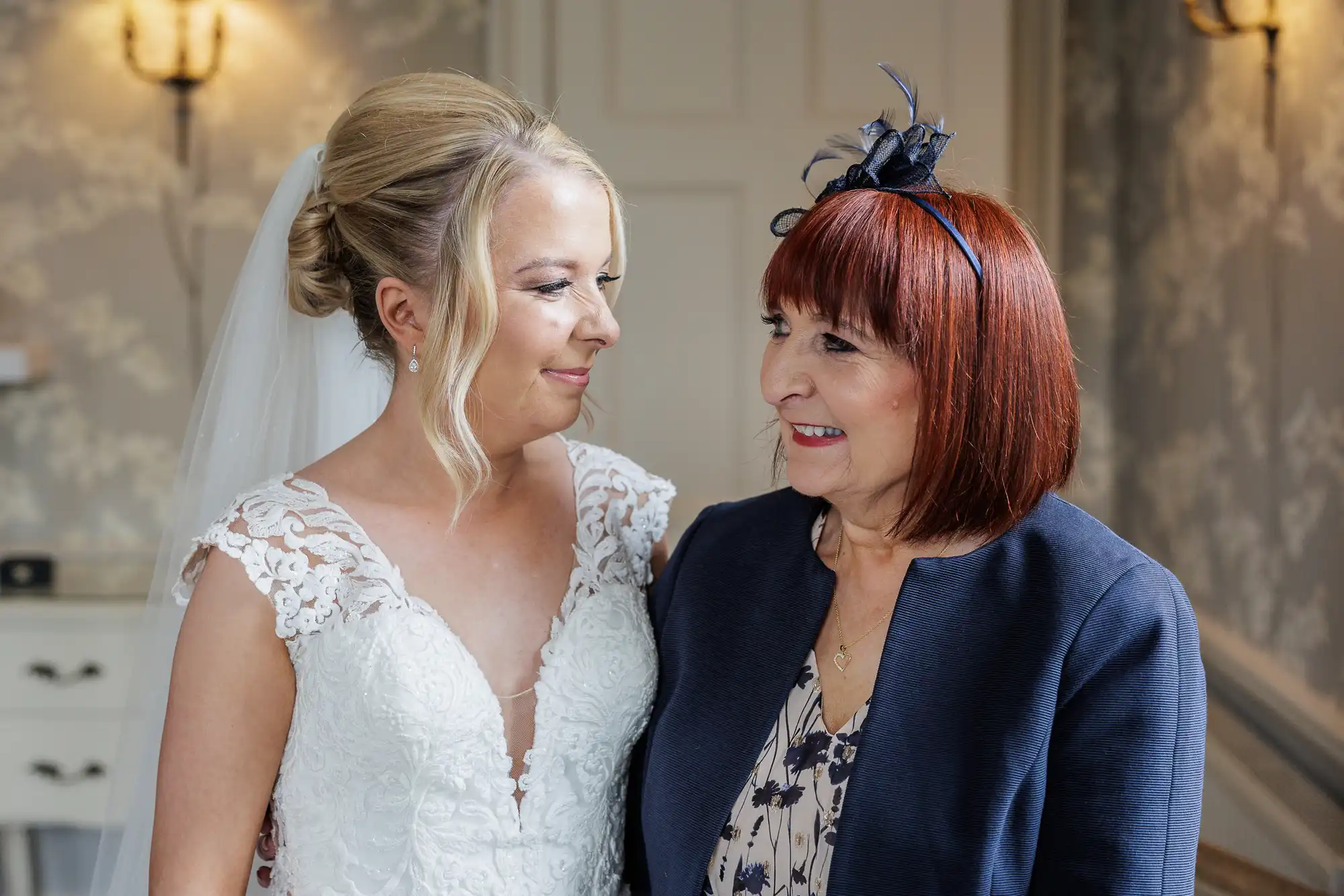 A bride in a white lace wedding dress and veil smiles warmly at an older woman with red hair wearing a navy jacket and fascinator; they stand close together indoors, facing each other with visible affection.