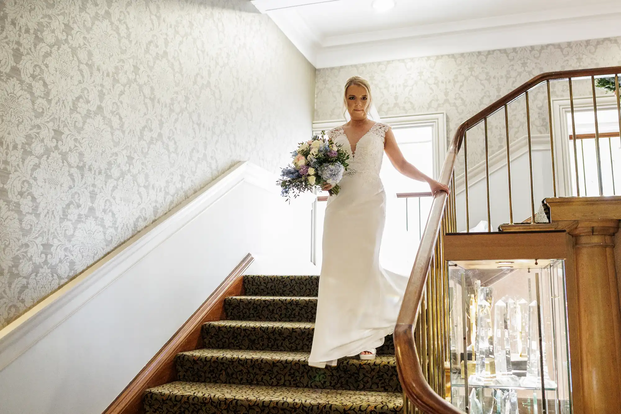 A bride in a white gown and holding a large bouquet descends a carpeted staircase with a wooden bannister in a decorated interior. She looks ahead, framed by patterned wallpaper and soft lighting.