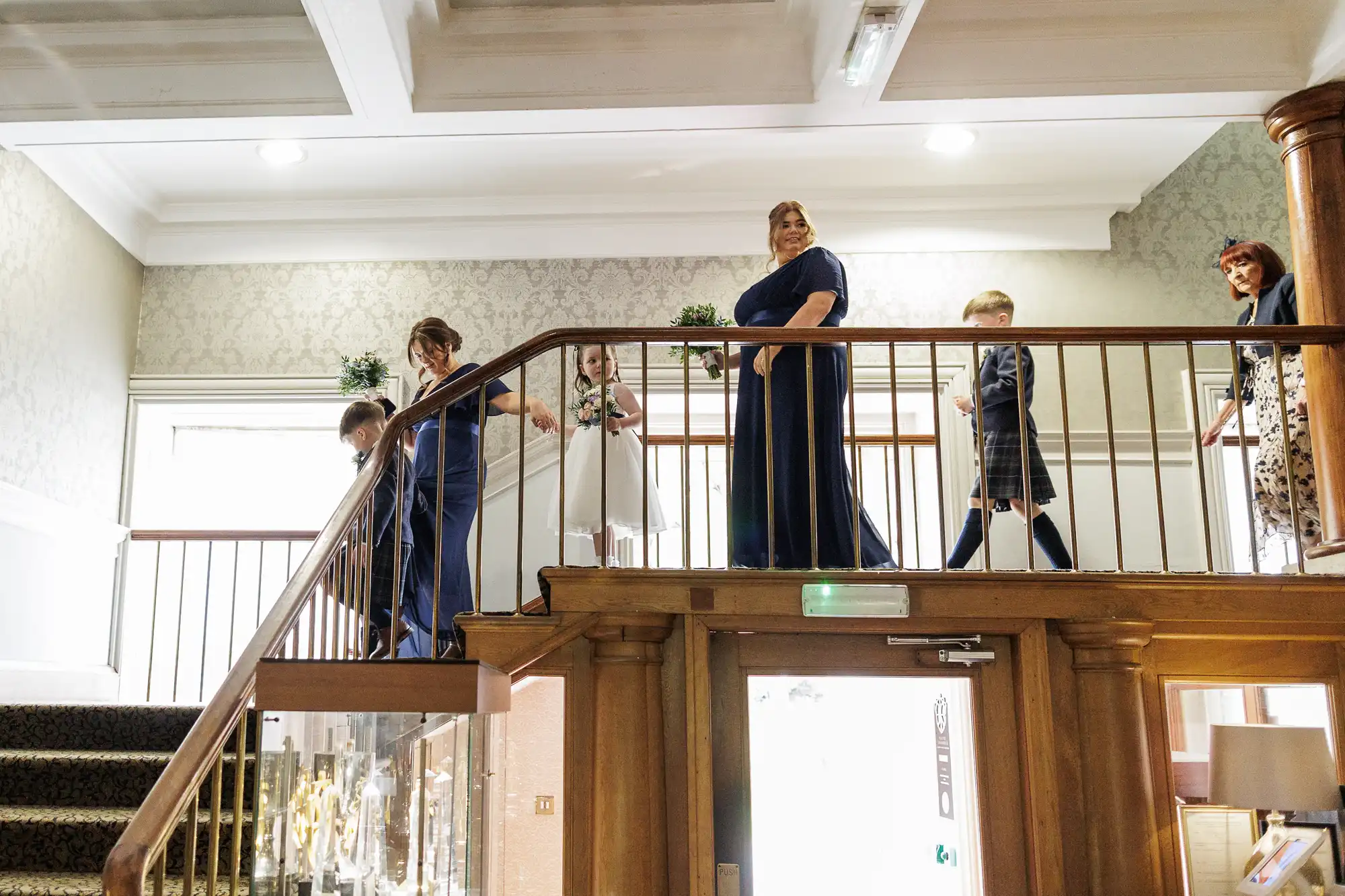 A group of people dressed formally walk across an indoor balcony. Three women in blue dresses and two children, one in white and one in check, move single file. The setting is elegant, with patterned wallpaper and wooden details.