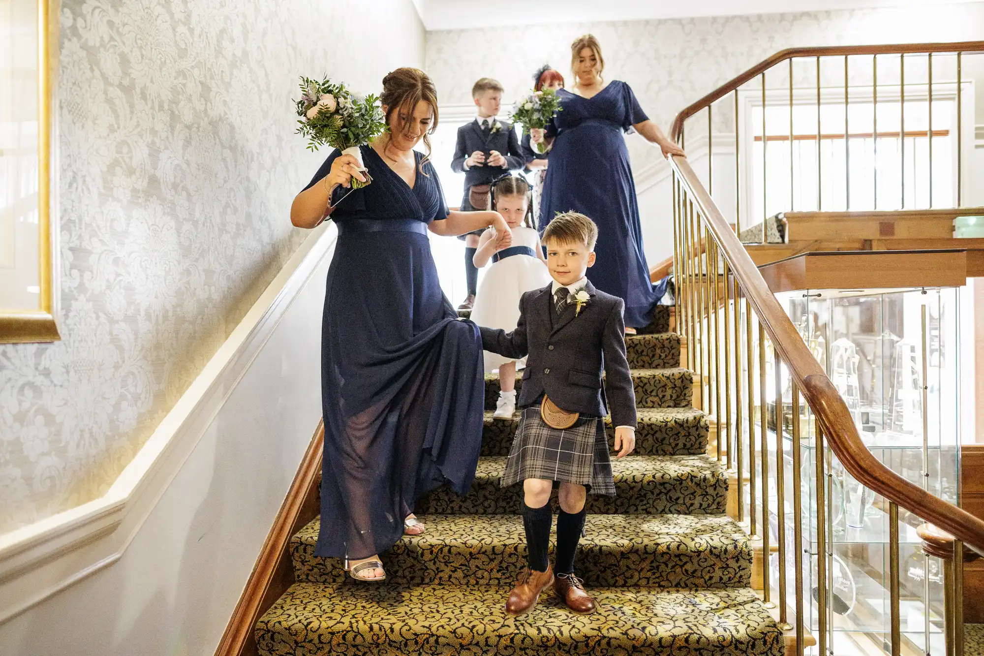 A group descends a carpeted staircase at a wedding: two women in blue dresses holding bouquets, two young boys in kilts and jackets, and one young girl in a white dress, all appearing cheerful and formal. The bannister is wooden with gold railings.