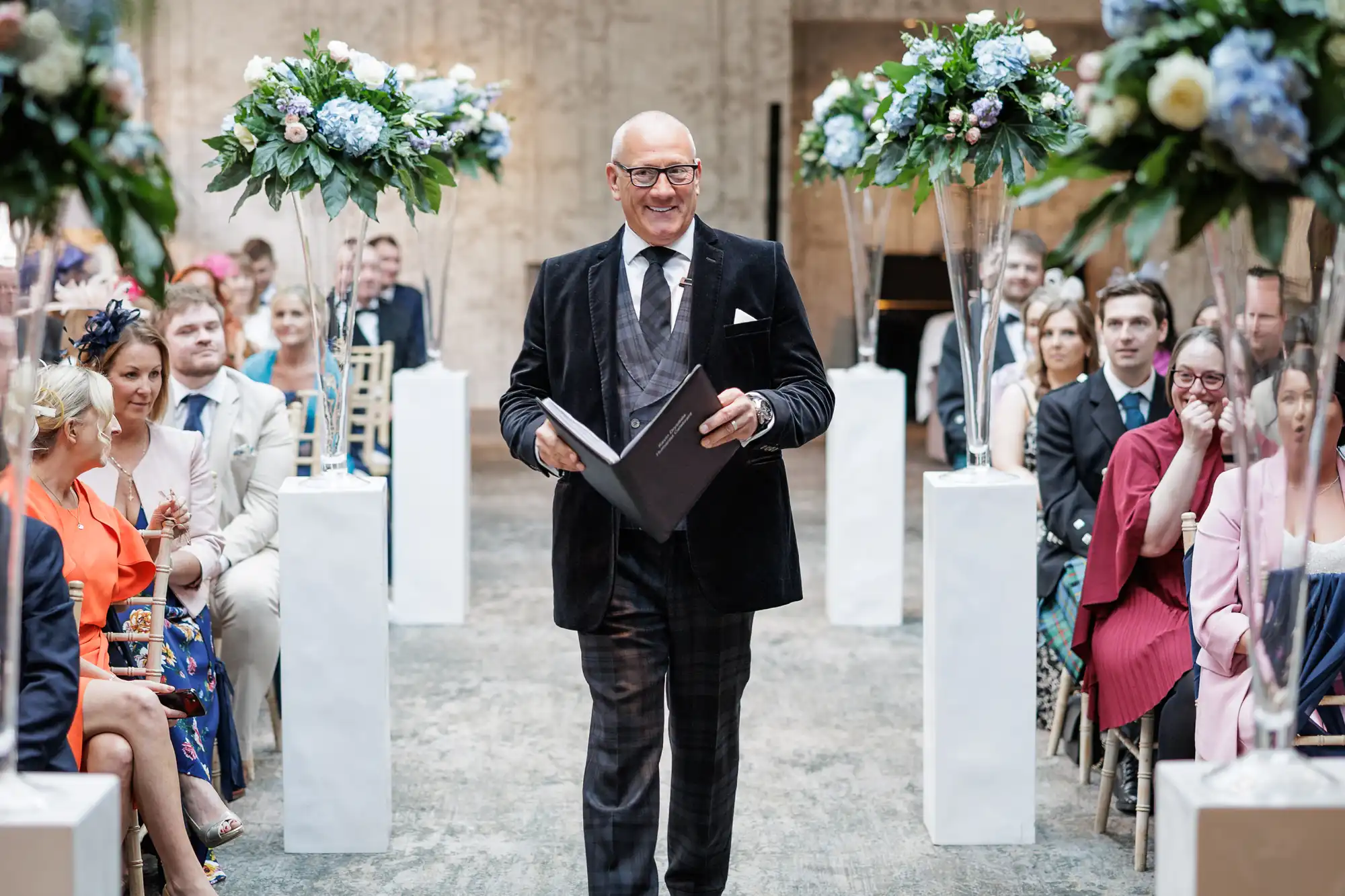 A smiling man in a black suit, spectacles, and holding a book walks down an indoor aisle lined with tall white flower arrangements. Seated guests in formal attire watch him, and the background shows neutral stone walls and soft lighting.