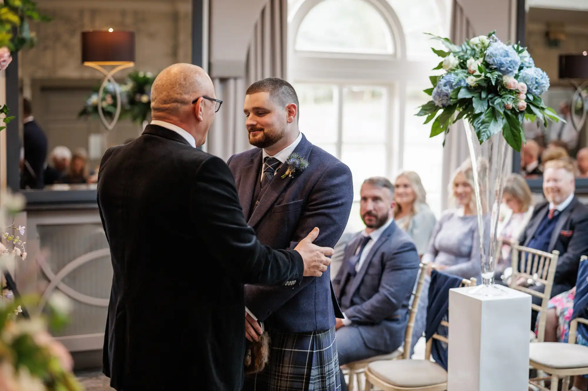 A man in a suit warmly greets a groom in a tartan kilt at a wedding ceremony. Guests are seated and watching. The room is decorated with blue and white flowers in tall vases and large windows let in natural light.