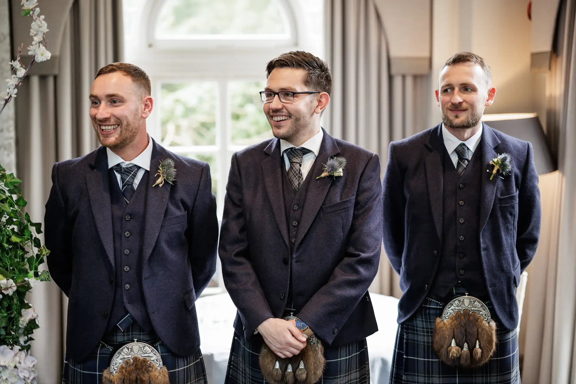 Three men stand indoors, smiling and dressed in traditional Scottish attire, including dark jackets, tartan kilts, and sporrans. They wear ties and buttonholes, with a window and light curtains in the background, giving a formal and festive atmosphere.