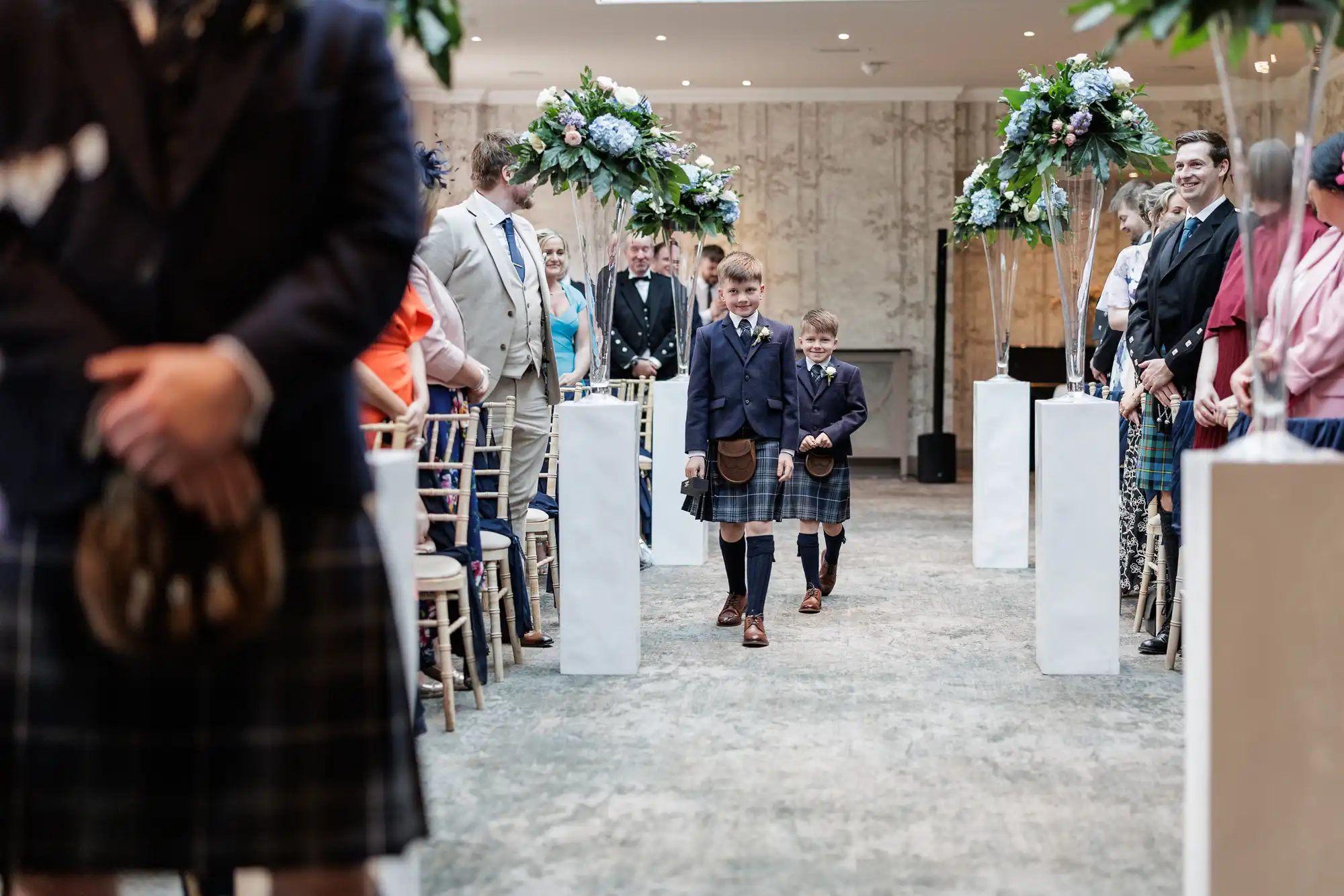 Two young boys in kilts and blazers walk down an aisle at a formal indoor event, possibly a wedding. Guests, some also in kilts, are seated on either side. Tall white stands with green and blue floral arrangements line the aisle.