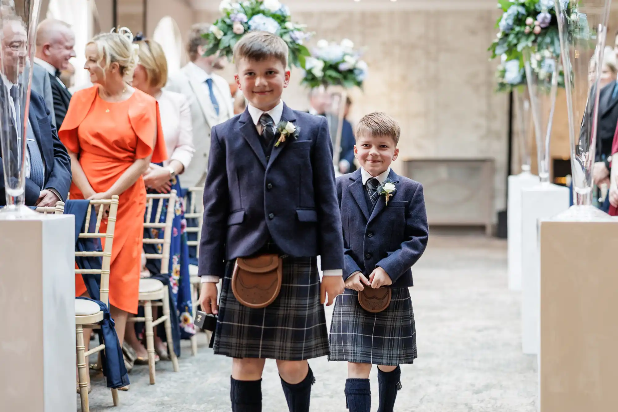 Two young boys in kilts, knee-high socks, and navy blazers walk down an aisle at a formal indoor event. Guests, some seated and some standing, watch them. The venue is decorated with floral arrangements and white pillars. The mood is festive and celebratory.