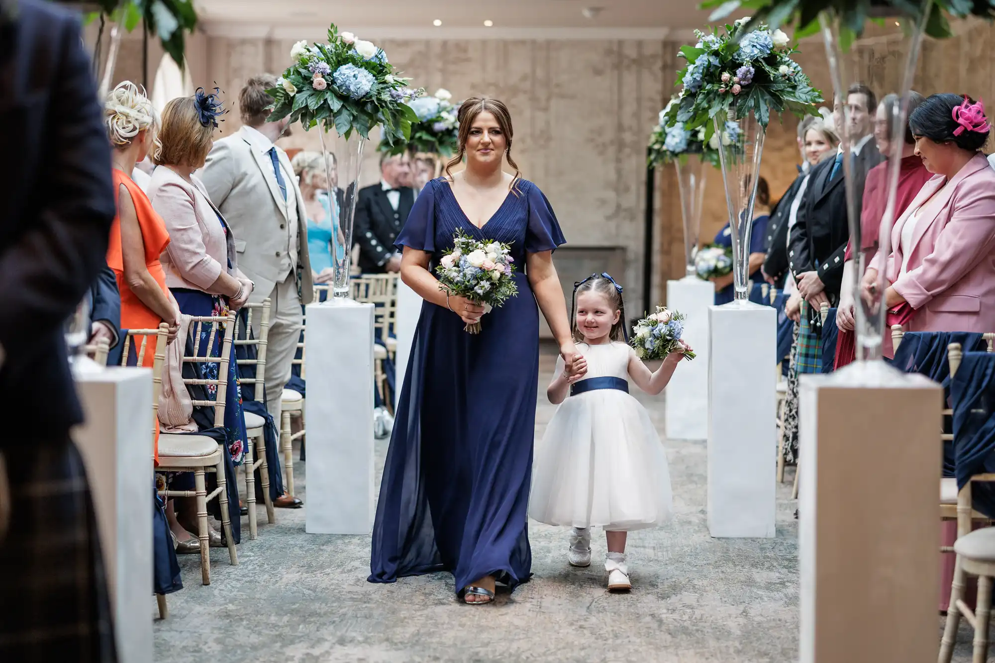 A woman in a navy blue dress walks down an aisle with a smiling young girl in a white dress and navy sash. Both hold flower bouquets. Guests in formal attire sit on either side. Tall floral arrangements decorate the aisle. The setting is bright and elegant.