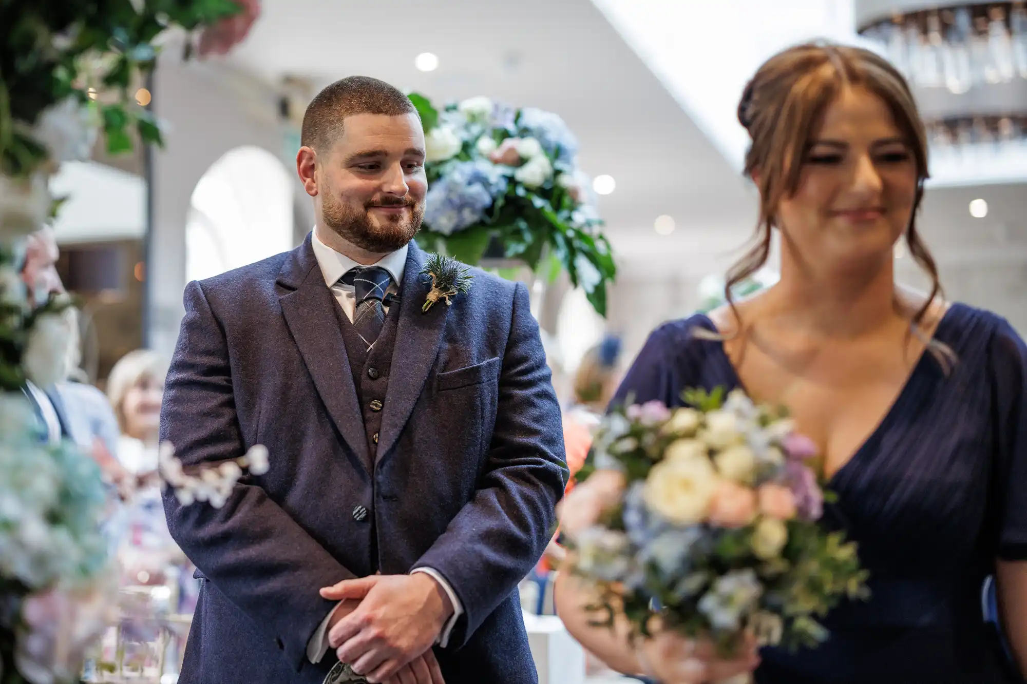 A man in a navy suit with a buttonhole smiles and looks to his right, standing indoors at a formal event. In the foreground, a woman in a blue dress holds a bunch of flowers. The background is softly focused, showing floral decorations.