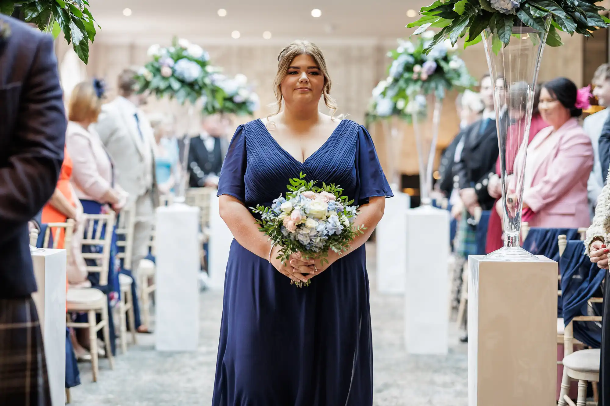 A woman in a navy blue dress stands in an aisle holding a bouquet of flowers. She faces the camera, with guests seated on both sides, some blurred. Tall floral arrangements decorate the aisle, and the setting appears to be an indoor wedding ceremony.