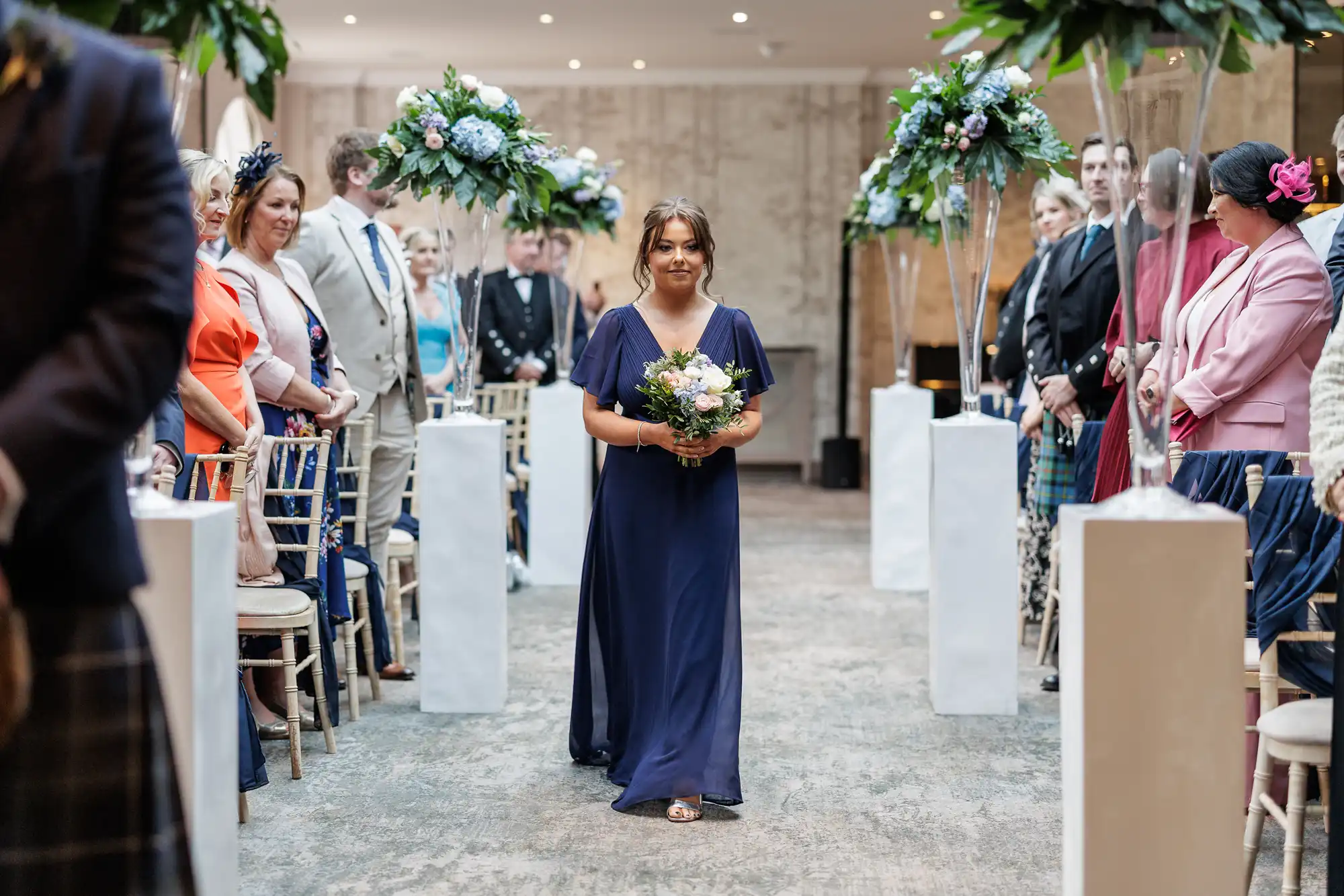 A bridesmaid in a navy blue dress walks down an indoor aisle, holding a bouquet. Guests on both sides watch, dressed in formal attire and seated on chairs. Tall floral arrangements decorate the aisle, and the space is softly lit and elegant.